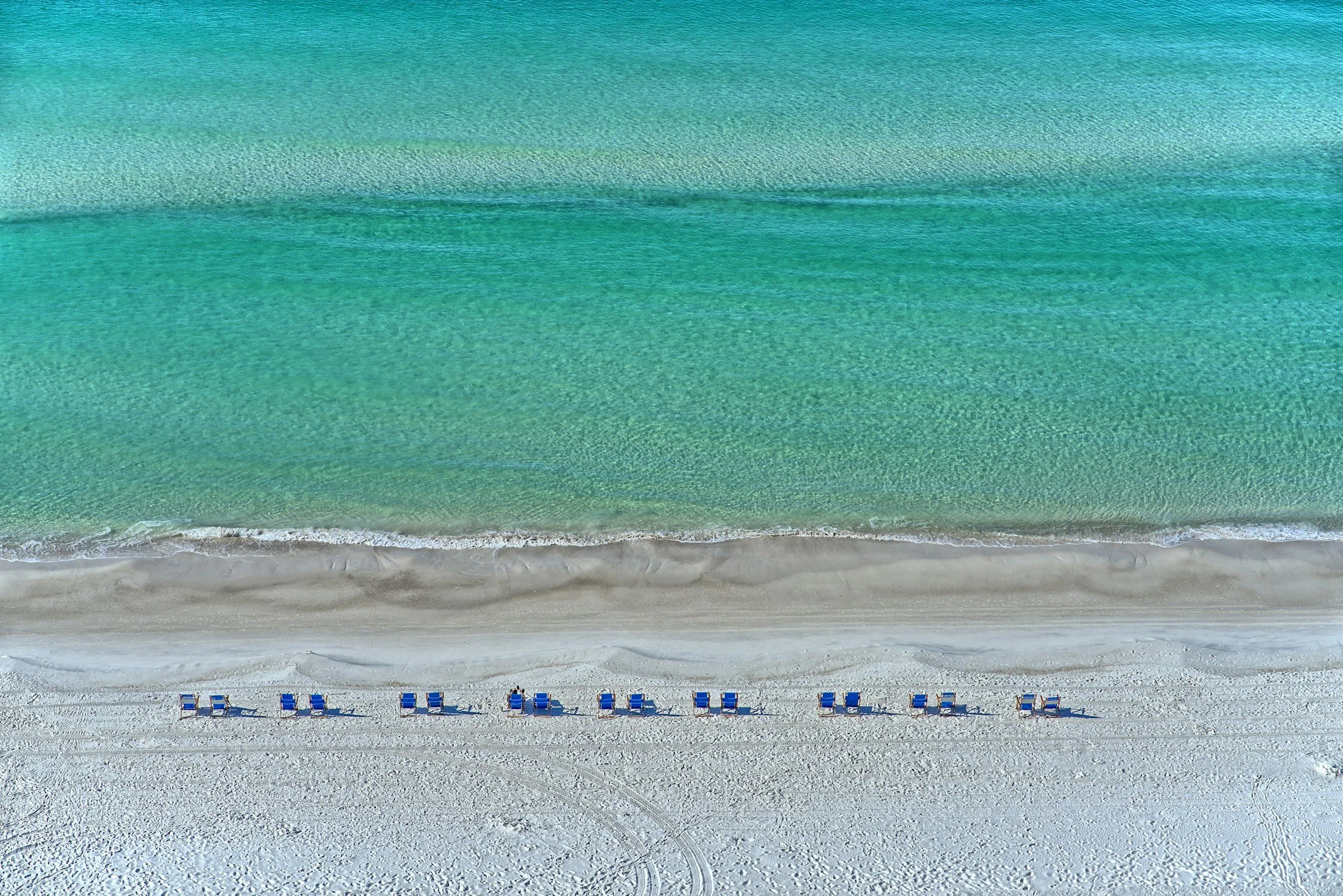 Aerial view of a beach with turquoise water and white sand, lined with blue lounge chairs and umbrellas.