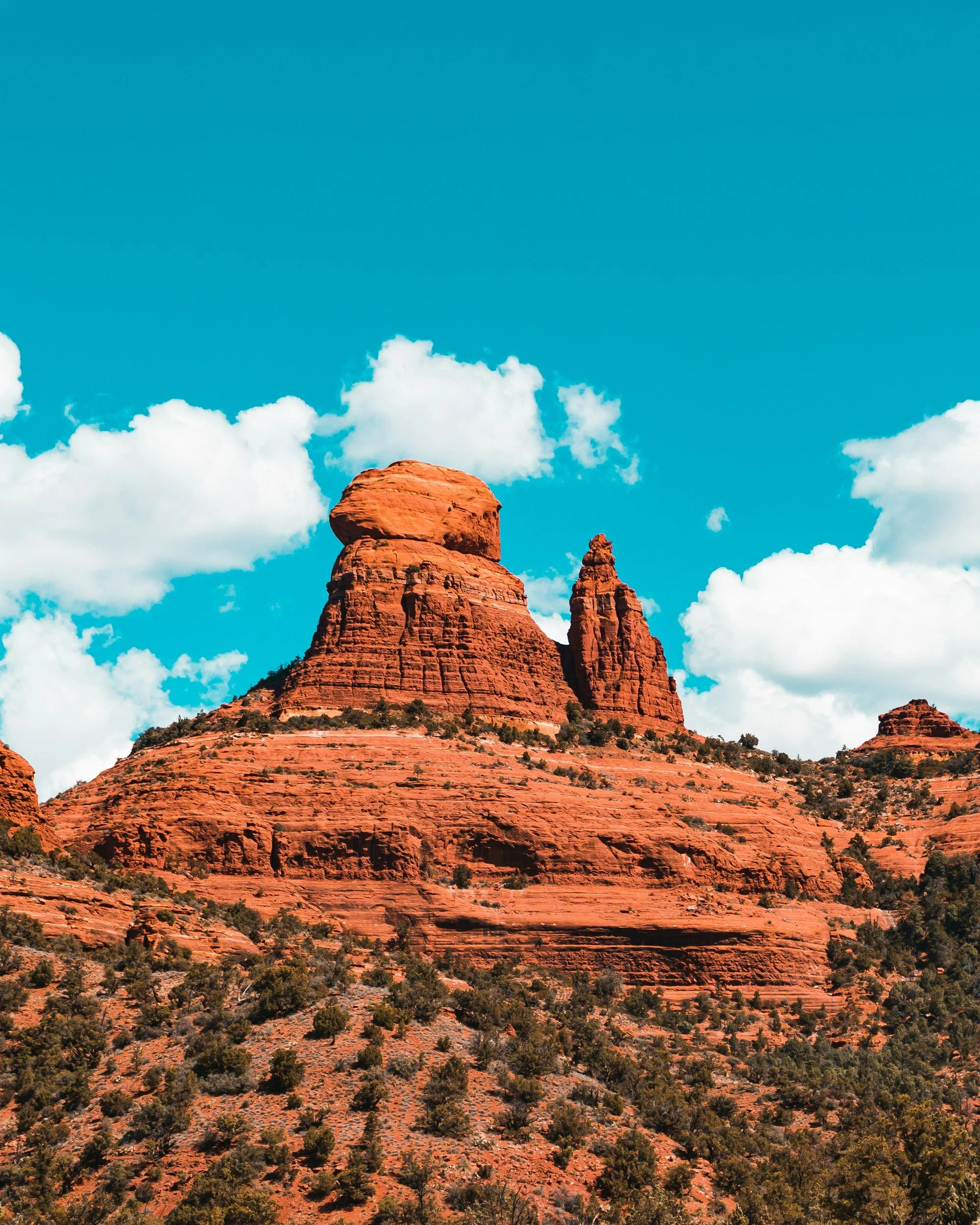 Sunset over red rock formations in a desert landscape with trees in the foreground and a partly cloudy sky