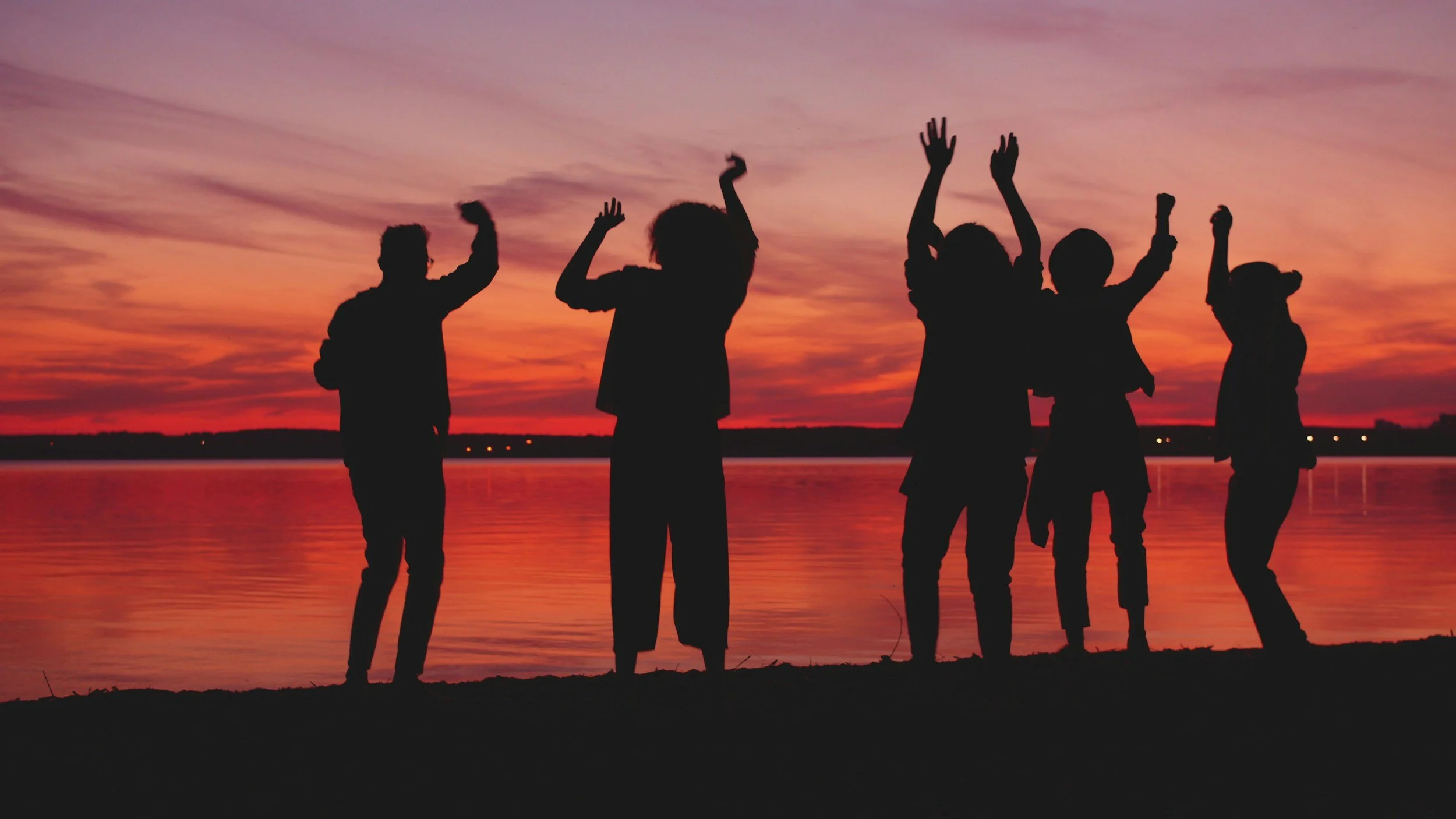 Silhouettes of five people standing by a body of water during a colorful sunset, with their arms raised and interacting with each other.