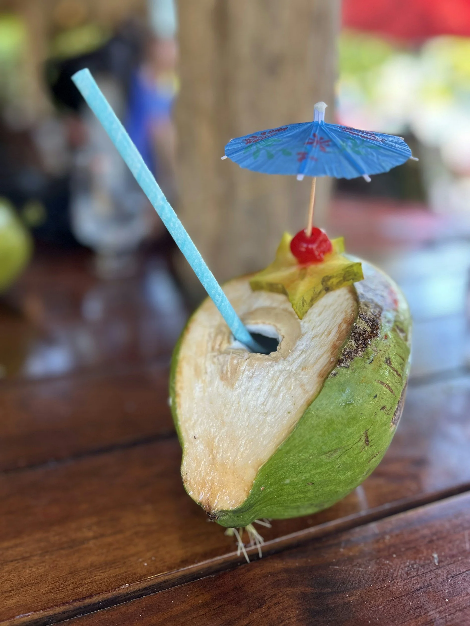 A tropical drink served in a hollowed-out coconut with a blue paper umbrella and a blue straw, garnished with a cherry, on a wooden table.