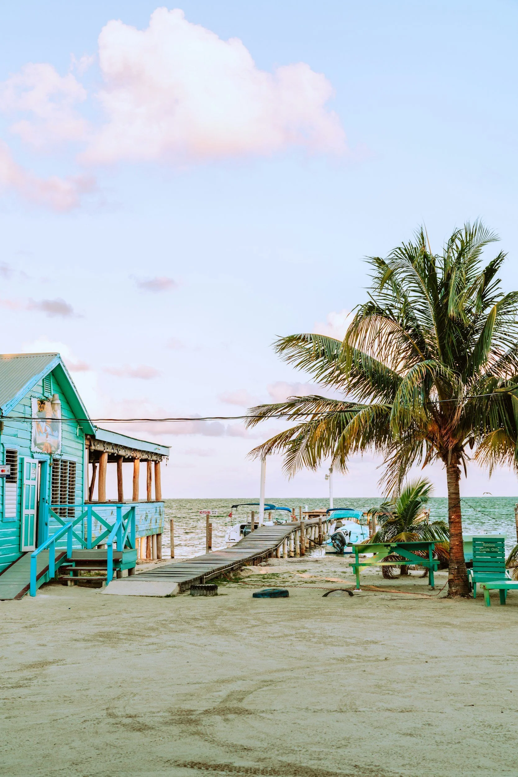A beach scene with a wooden pier, colorful beach house, palm tree, and boats by the water, under a partly cloudy sky.