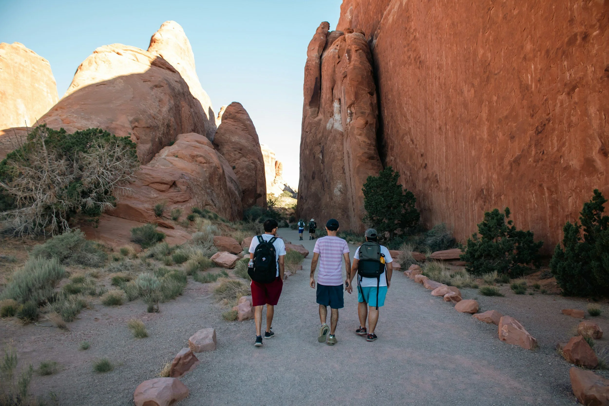 A group of people hiking through a desert landscape with tall red rock formations and sparse desert vegetation.