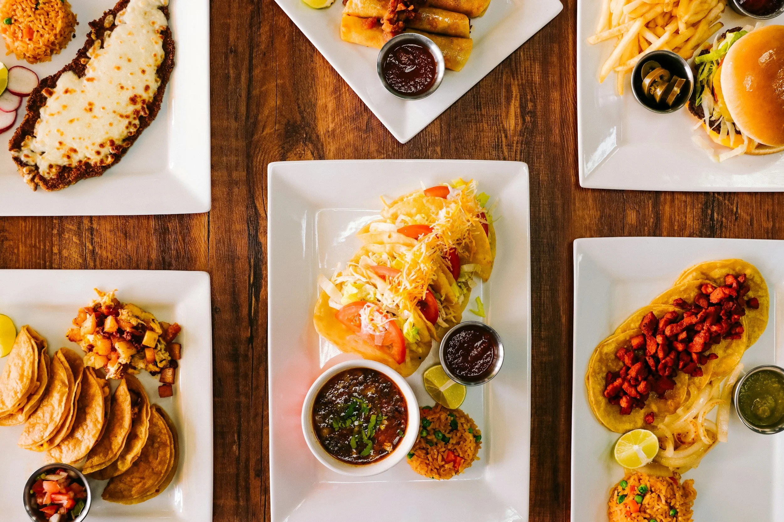 Assorted Mexican dishes on white plates, including tacos, tamales, and Mexican rice, with small bowls of salsa and lime wedges, arranged on a wooden table.
