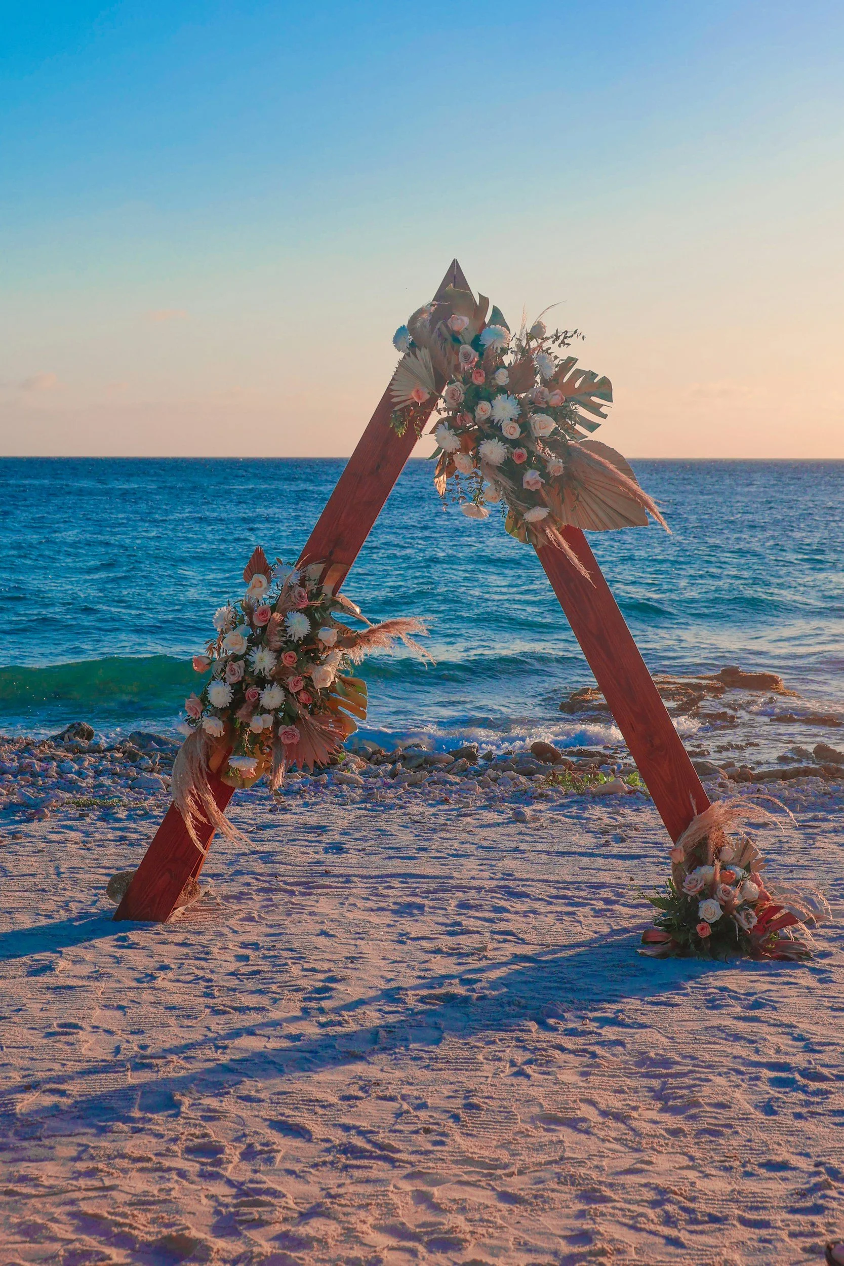 Decorative wooden triangle frame with pink and white flowers, set on a sandy beach with ocean and sunset in the background.