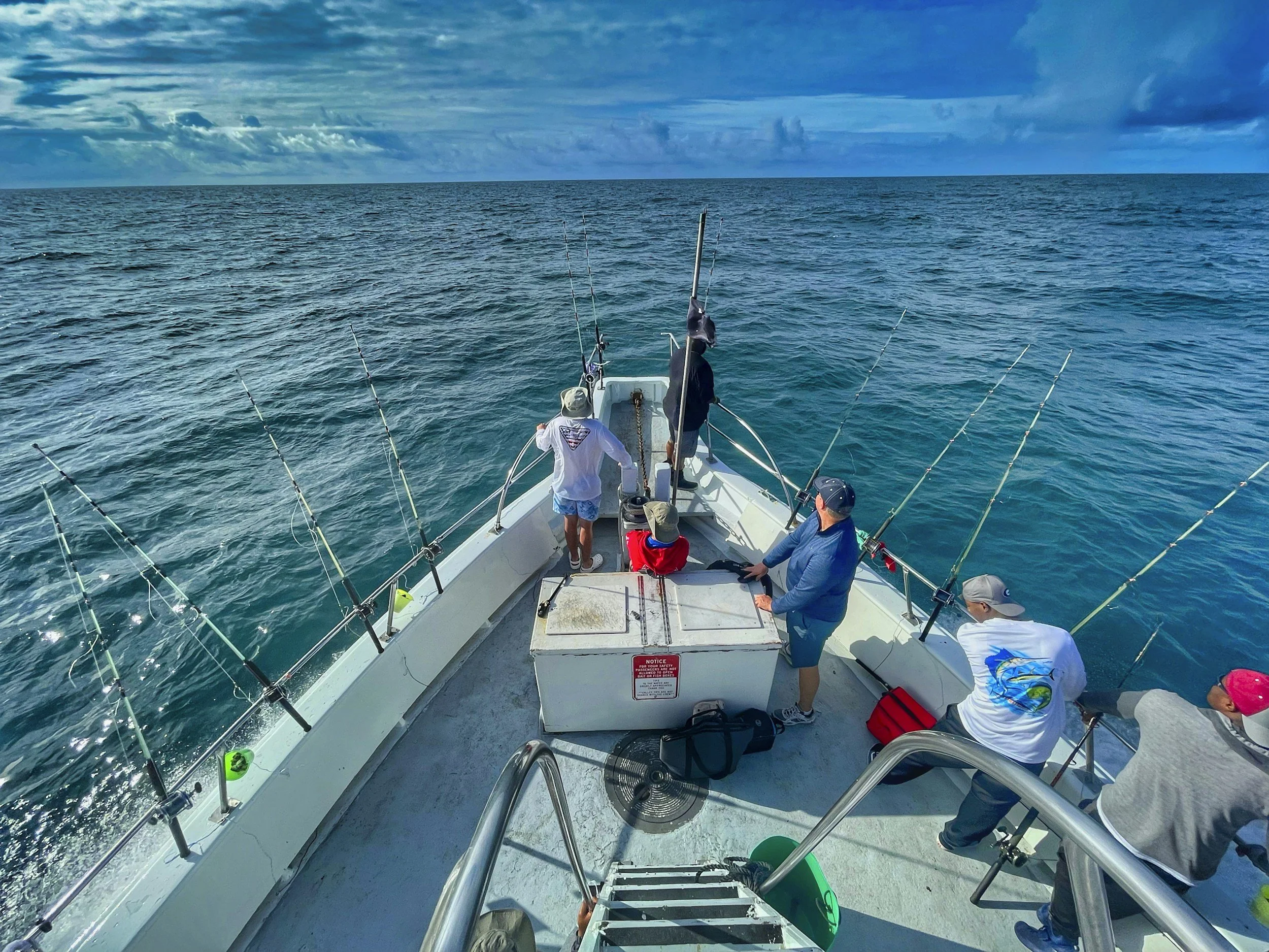 People fishing from a boat in the open ocean, with multiple fishing rods set up along the boat. The scene includes cloudy skies and calm blue waters.