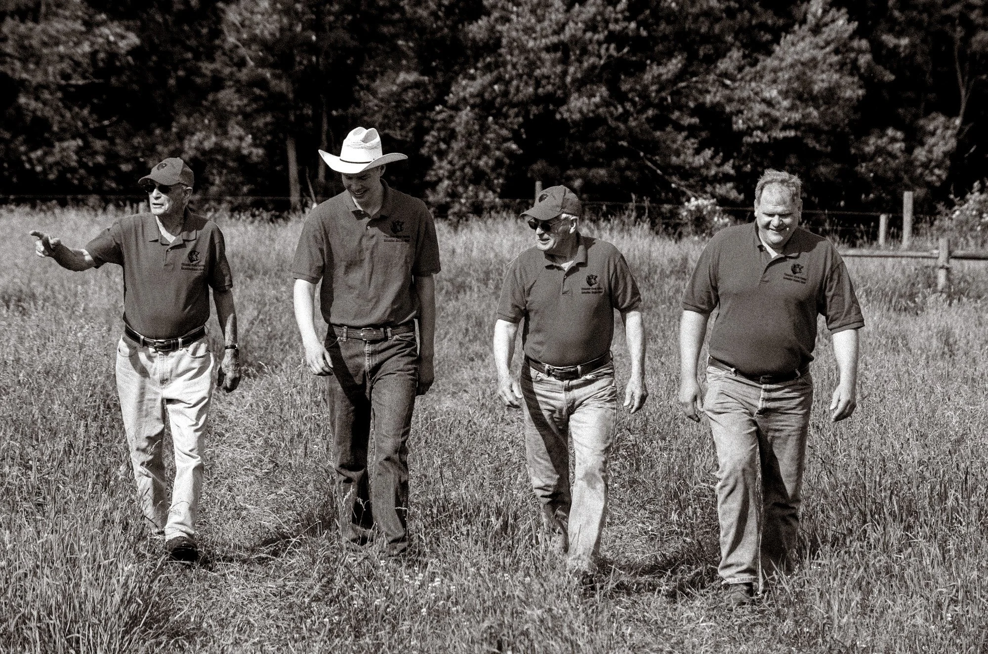 Four men walking through a grassy field, dressed casually in polo shirts and jeans, with one wearing a cowboy hat, in a rural outdoor setting with trees in the background.