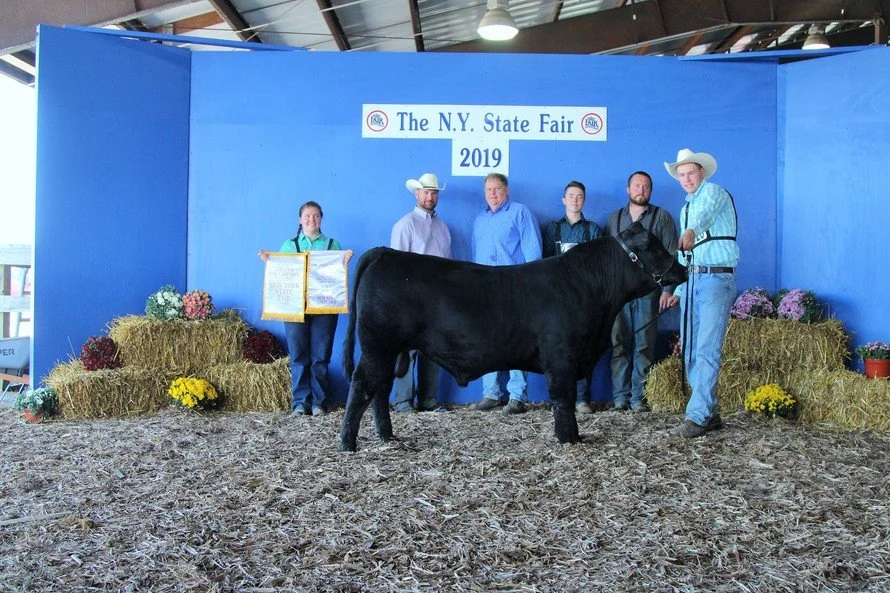 Group of people posing with a black calf and awards at the 2019 New York State Fair, with hay bales and flowers as decorations.