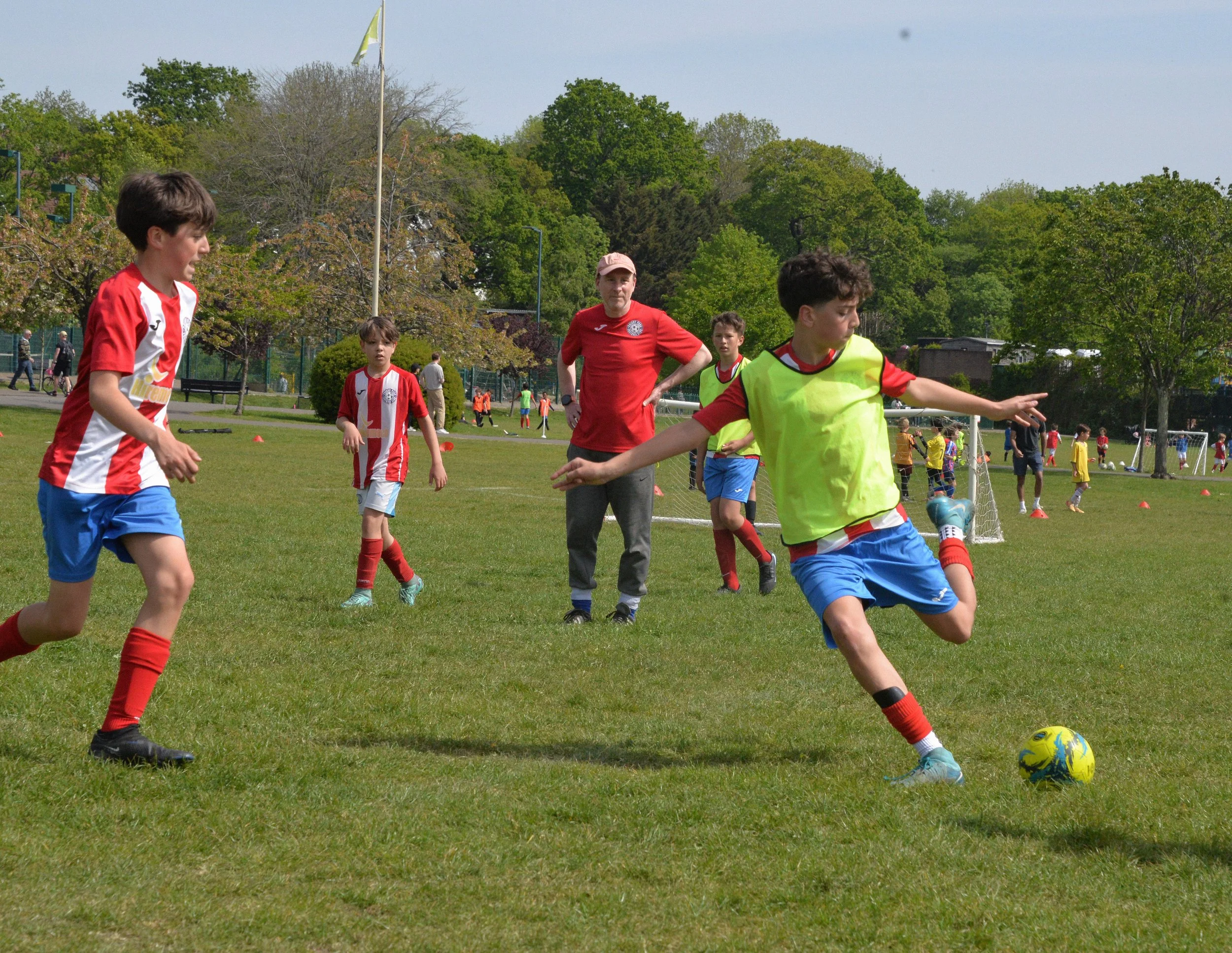 Children playing soccer on a grassy field with a coach or adult supervising in the background. The children are wearing sports uniforms, some in neon yellow bibs, and are engaged in a soccer activity.