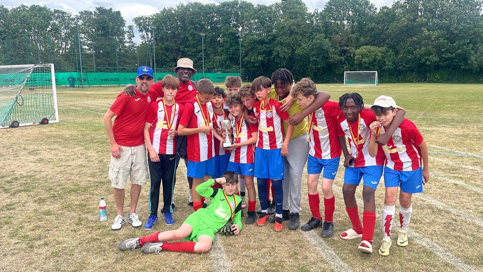 A group of young soccer players and two coaches celebrating with a trophy on a soccer field. The team is dressed in red and white uniforms, and one player is lying on the ground in a bright green goalkeeper uniform. The background shows soccer goals 