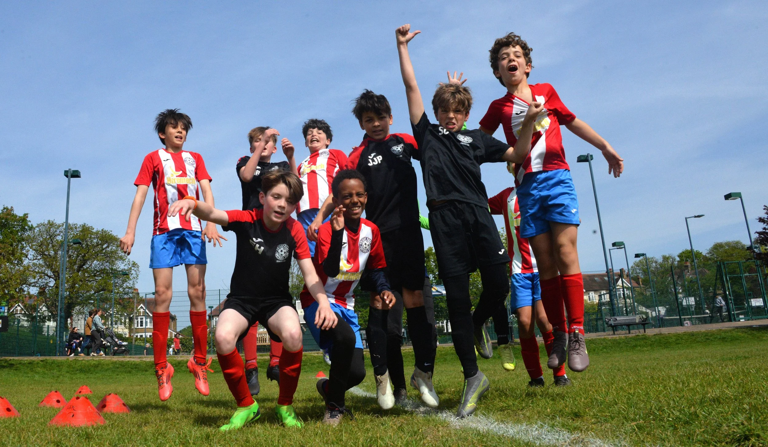 Group of young boys playing soccer on a field, some jumping in the air, wearing sports uniforms, with orange cones on the ground and a blue sky overhead.