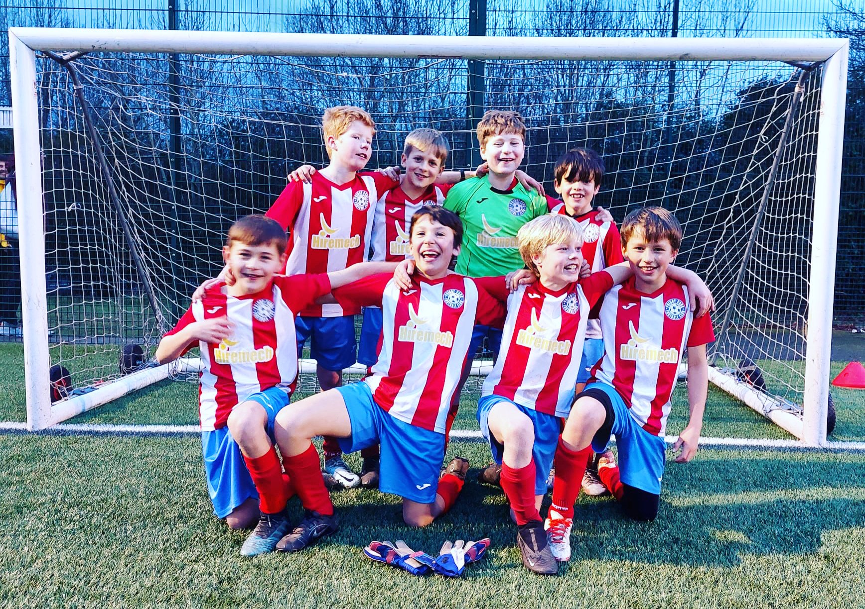 Young boys in soccer uniforms posing in front of a goal on a soccer field. They are smiling and appear happy, with some kneeling and others standing behind them. One of the boys in the back row is wearing a green goalkeeper jersey.