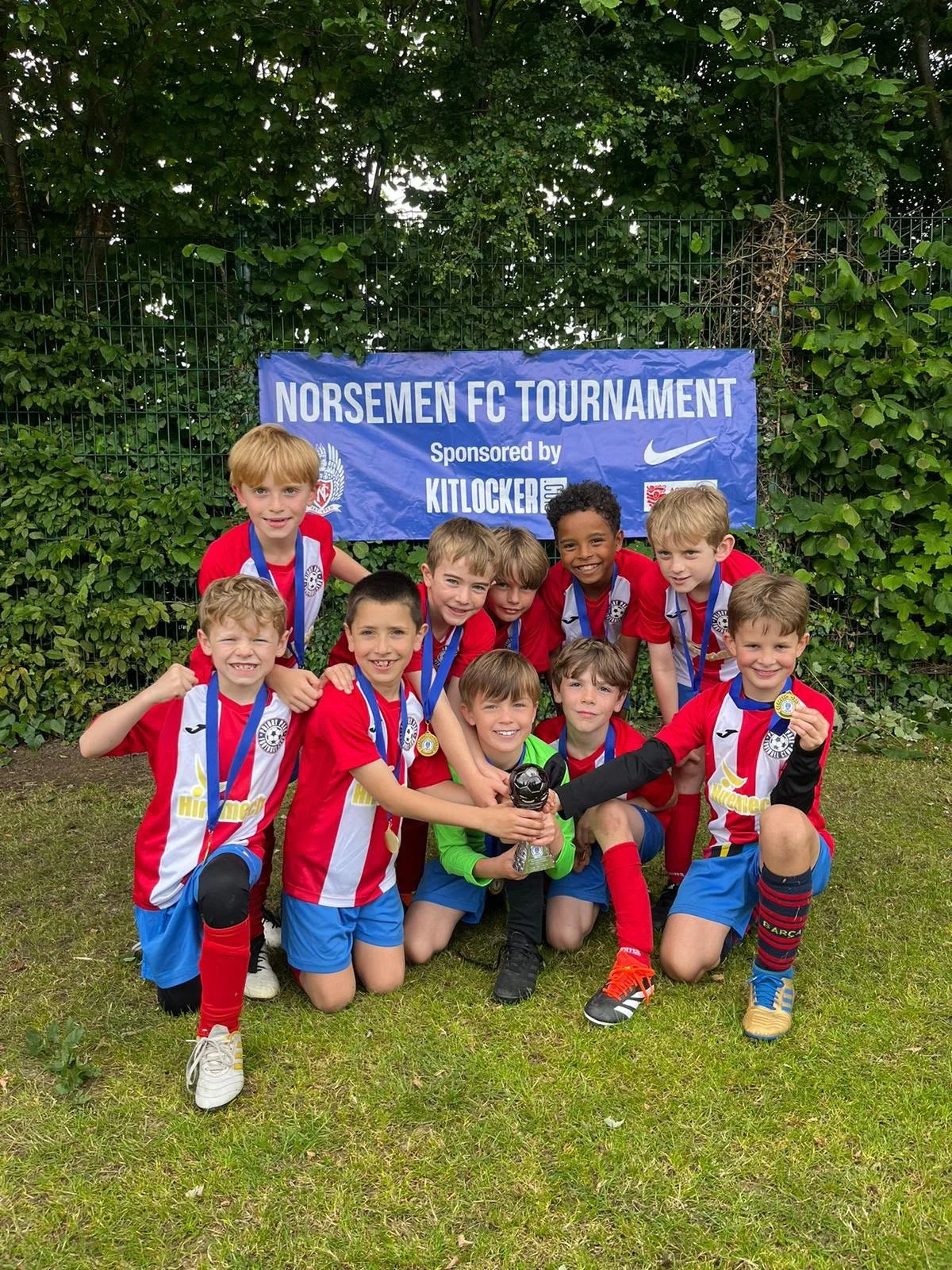 Youth soccer team in red and blue uniforms holding medals and a trophy, posing in front of a blue banner with 'Norsemen FC Tournament' written on it, surrounded by greenery.