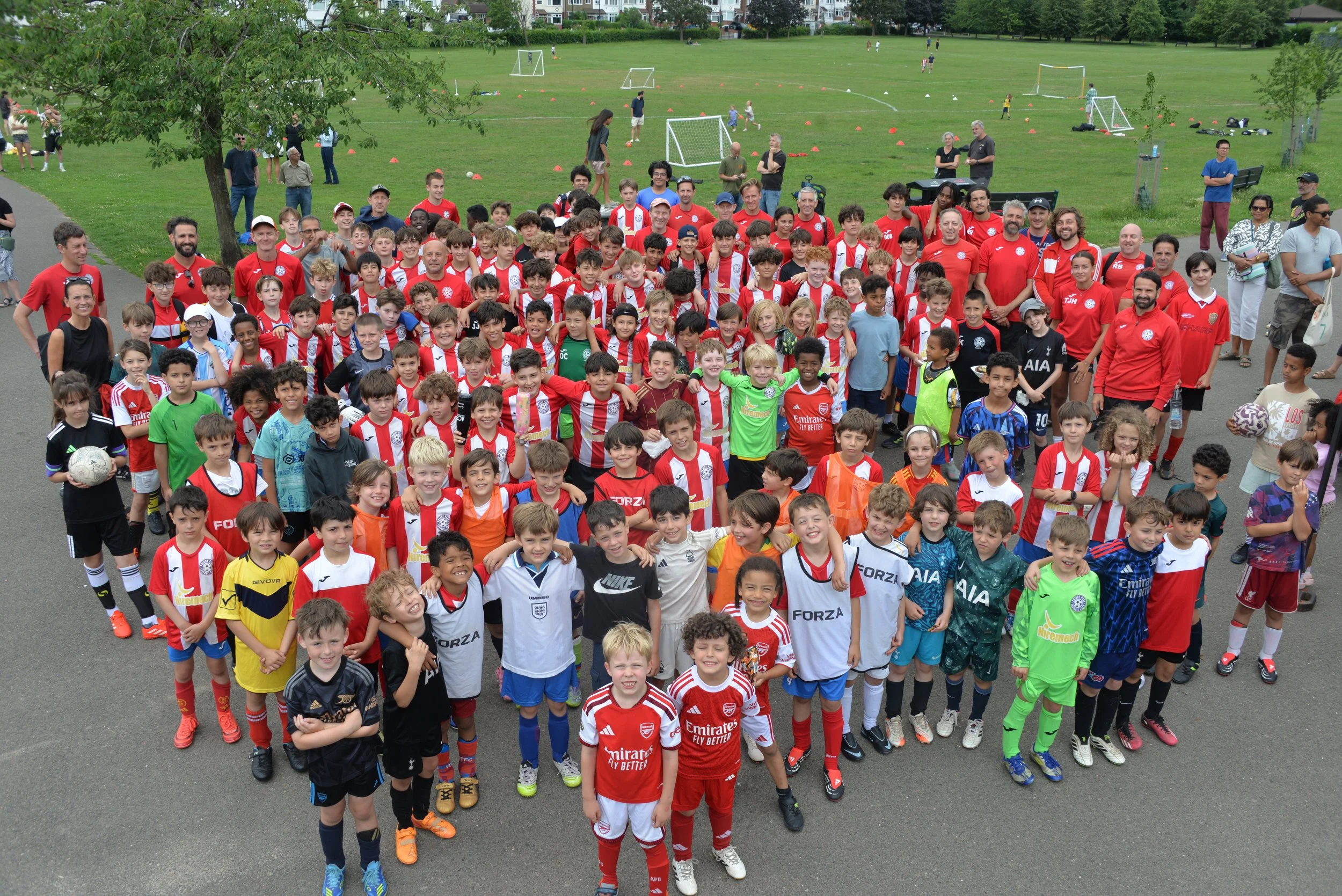A large group of children and adults gather outdoors on a cloudy day, wearing colorful sports jerseys and casual clothing, with a grassy park and soccer fields in the background.