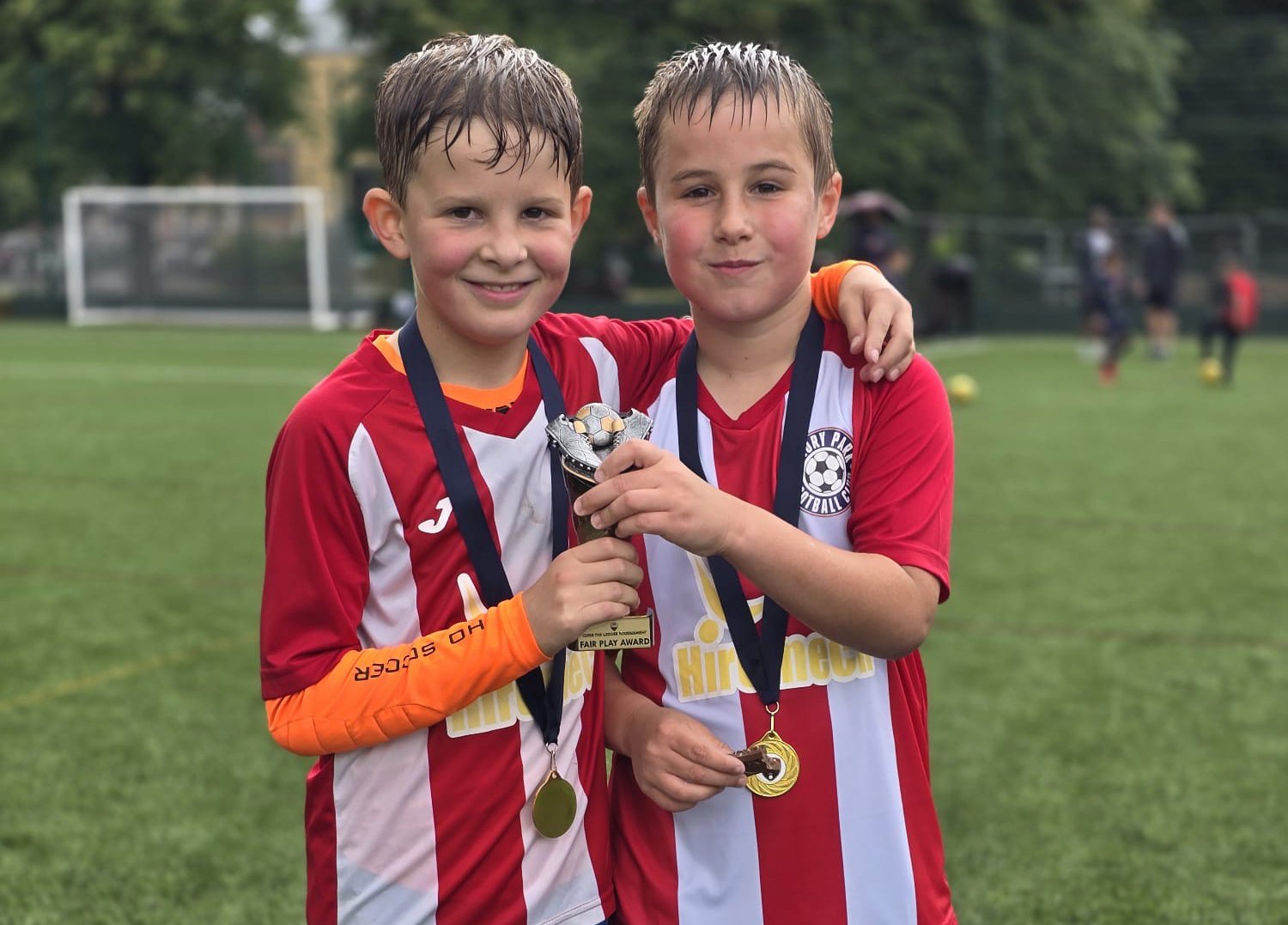 Two young boys in red and white soccer uniforms are smiling and standing on a soccer field, holding a trophy and medals after a game.