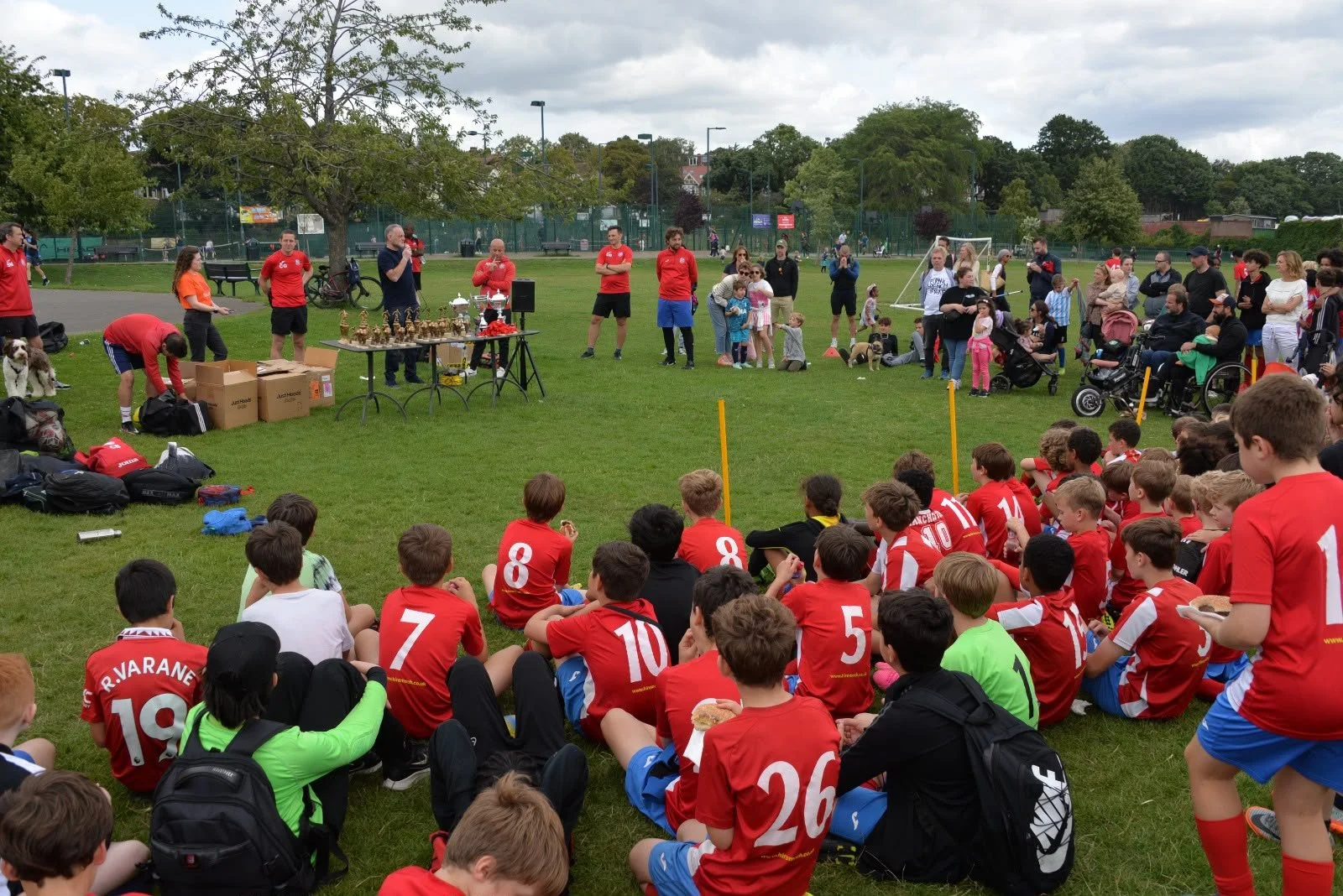 Children in red sports jerseys sitting on the grass in a park, attending an outdoor event with adults and trophies displayed on tables, amid trees and a cloudy sky.