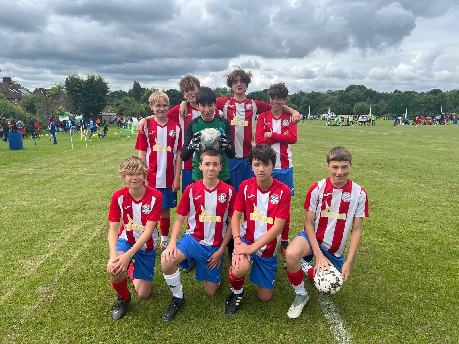 A group of young boys in soccer uniforms with red and white striped shirts and blue shorts, some kneeling and some standing, on a soccer field. They are posing for a team photo, with a goalkeeper holding a soccer ball in the middle. In the background