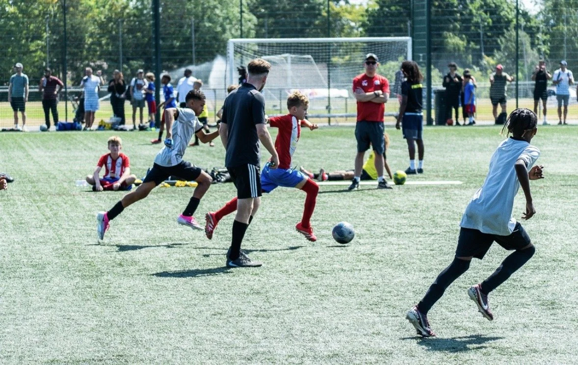 Children playing soccer on a field with spectators watching in the background.