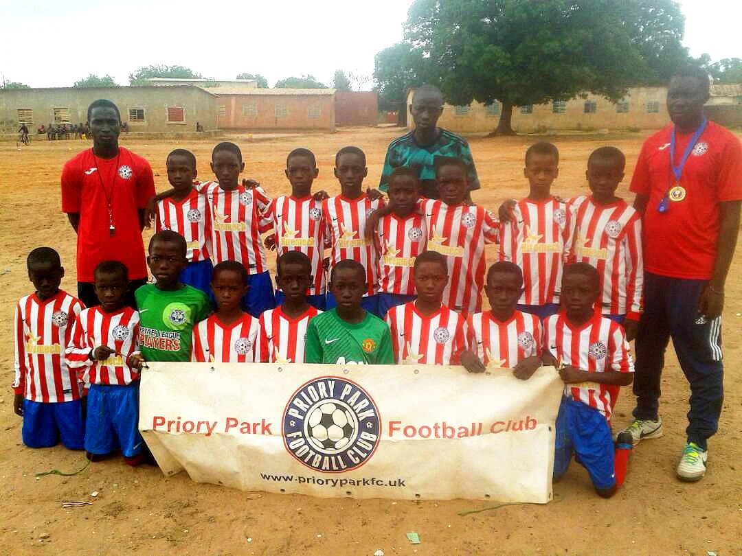 A youth soccer team from Priory Park Football Club posing on a soccer field with their coaches, holding a banner with the club's logo and website, wearing red and white striped jerseys and blue shorts.
