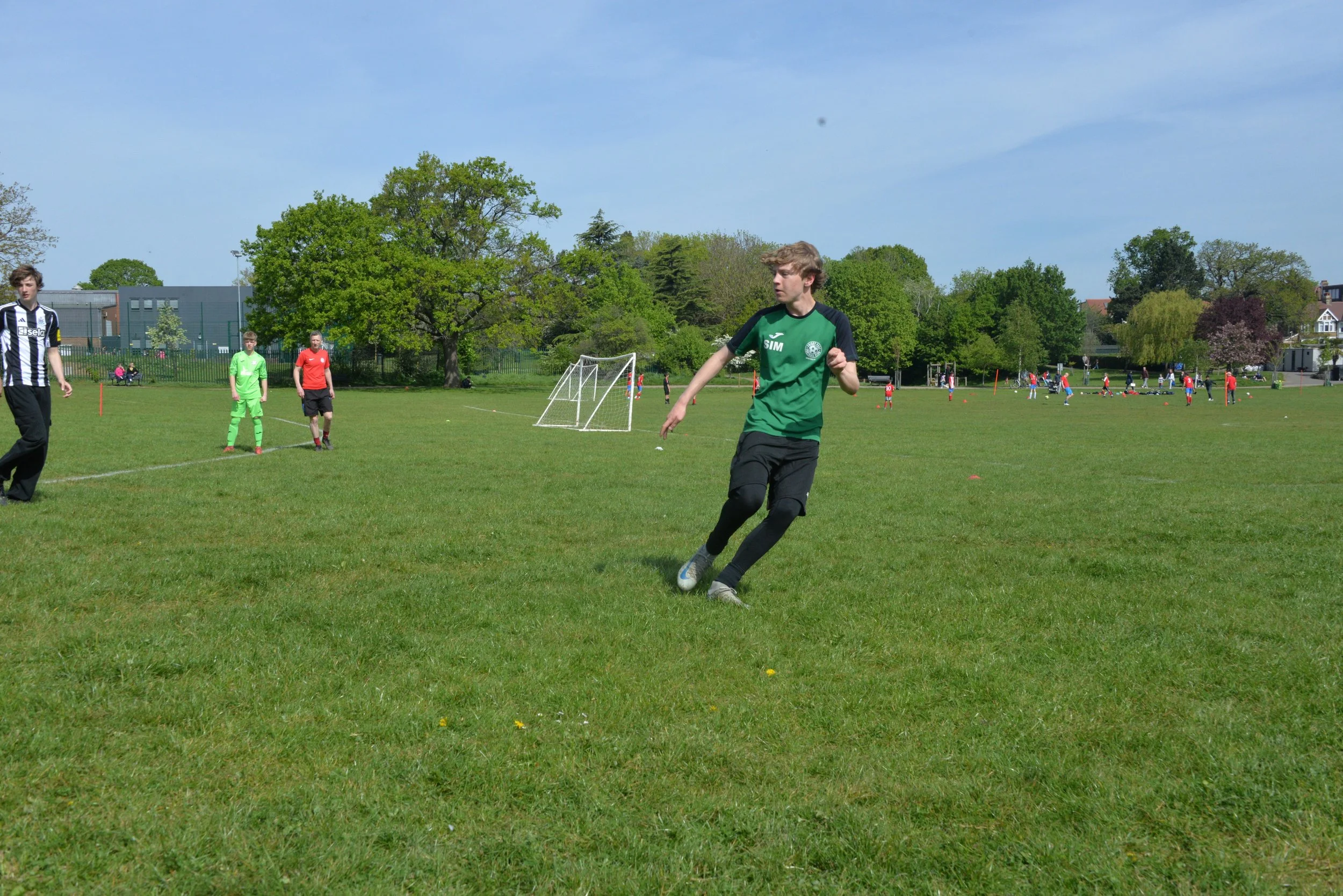 A group of young people playing soccer on a lush green field on a sunny day, with trees and buildings in the background.