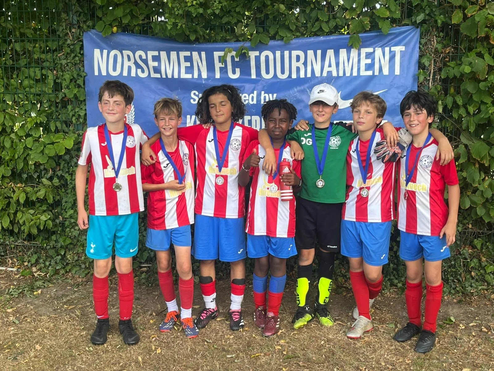 Group of young boys in soccer uniforms standing in front of a blue banner that reads 'Norsemen FC Tournament.' They are smiling, with some wearing medals around their necks. One boy is dressed as a goalkeeper in a green jersey and gloves.
