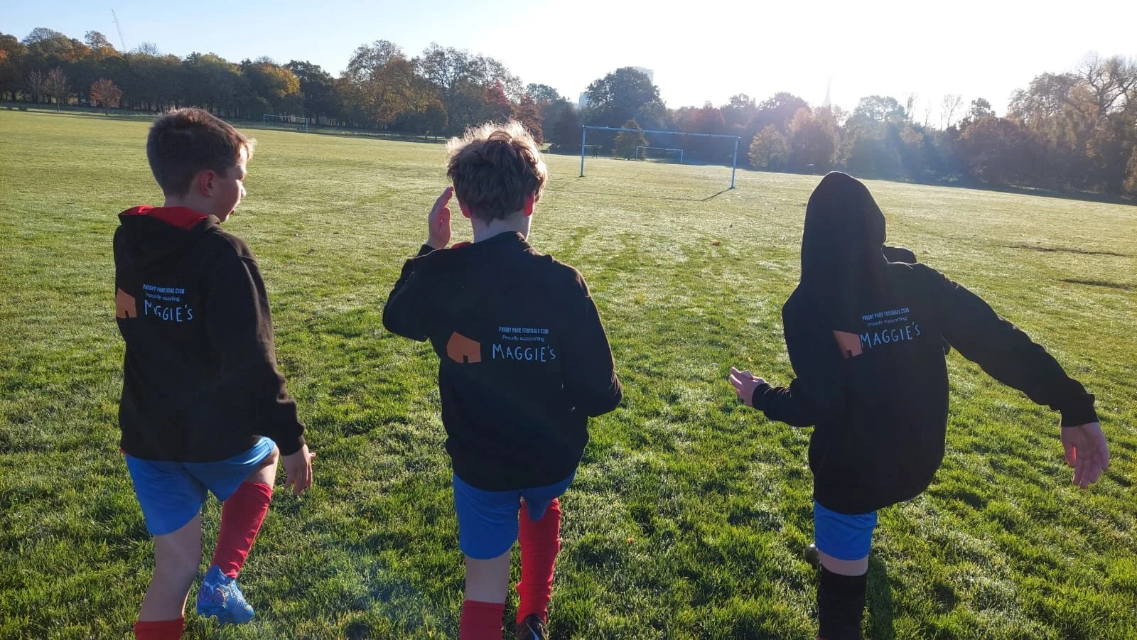 Three young boys wearing black hoodies with 'Maggie's' logo playing on a grassy field near a soccer goal during late afternoon.