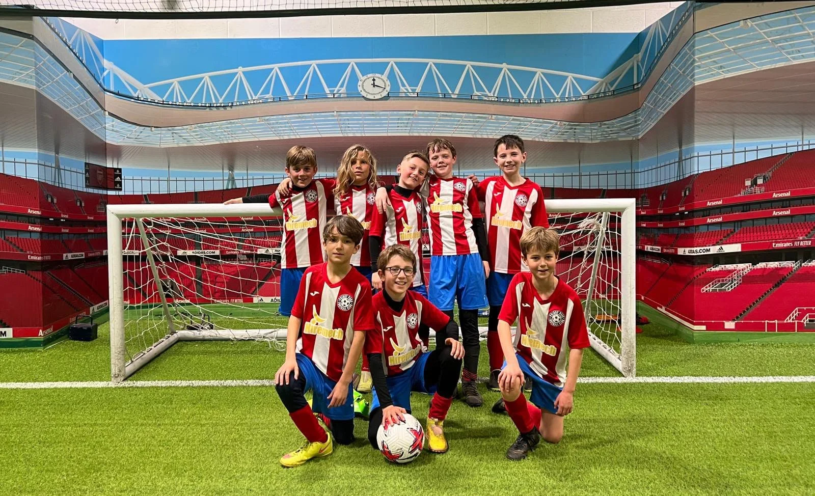A group of young boys and girls in soccer uniforms posing in front of a soccer goal inside a stadium. The children are smiling and appear to be part of a youth soccer team, with a soccer ball on the ground in front of them.