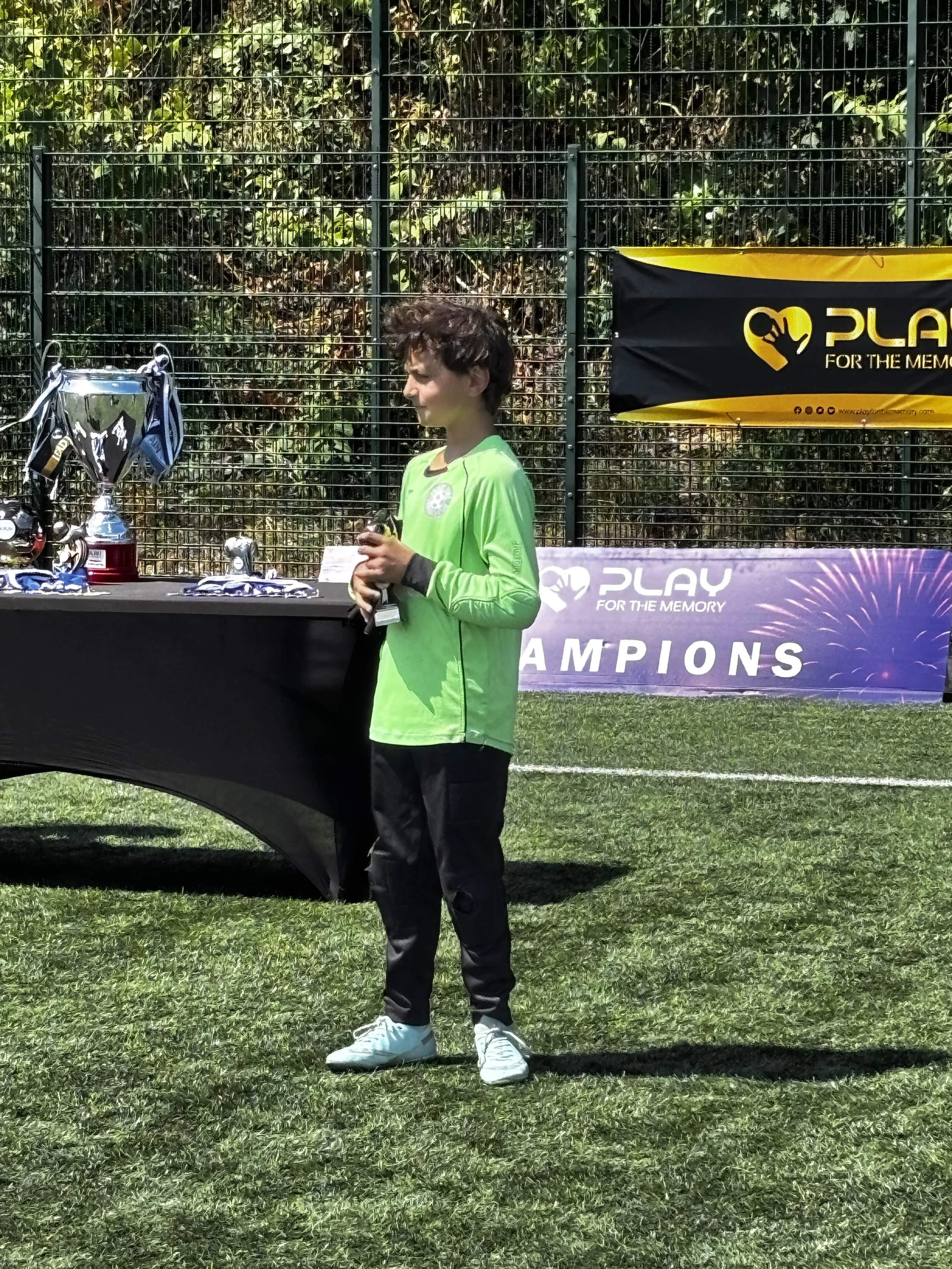 A young man standing on a grassy field wearing a bright green sports jersey, black pants, and white sneakers, holding a small object in his hand. Behind him, there is a table with trophies, medals, and sports gear, with a fence and greenery in the ba