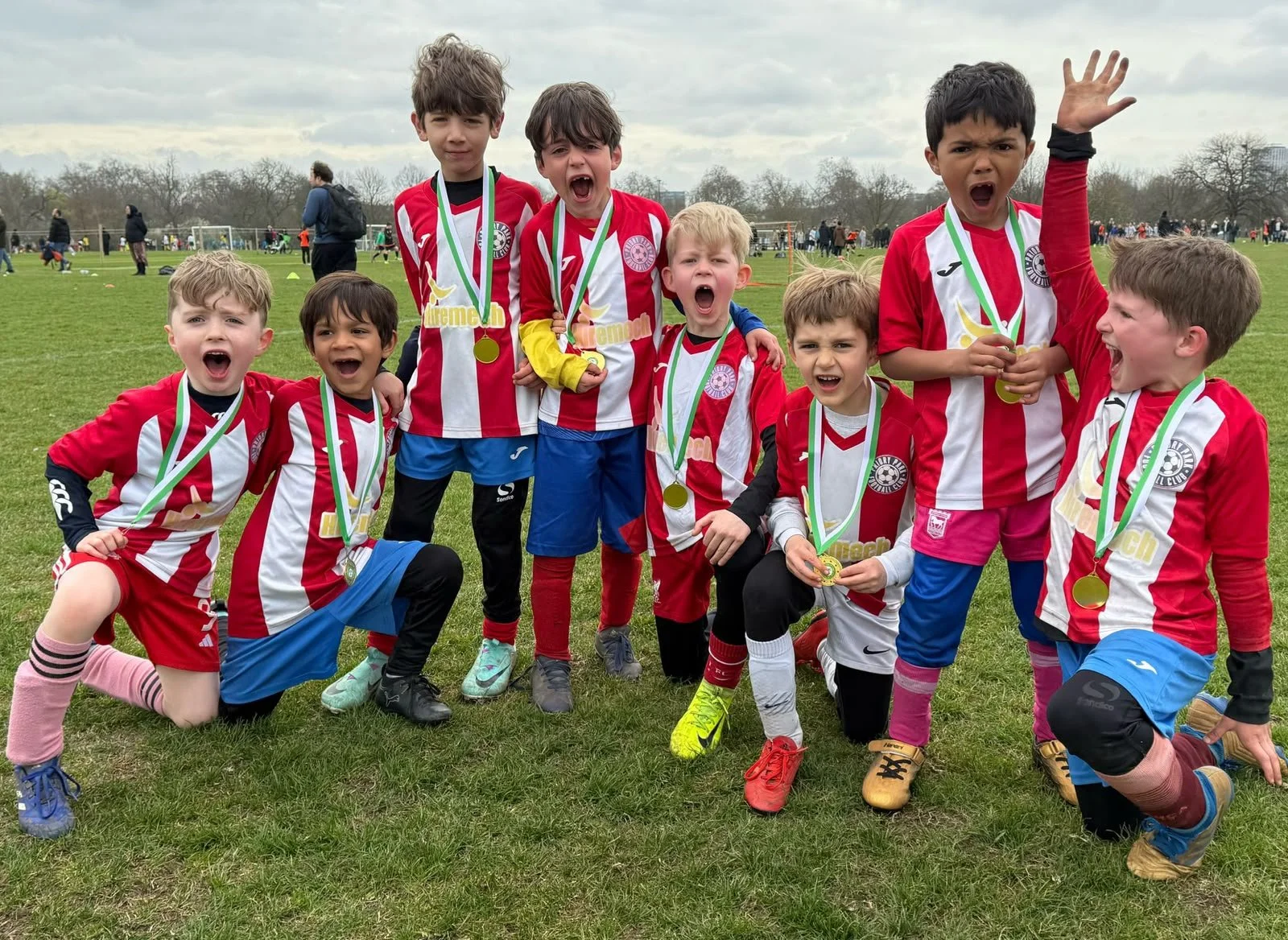 Young boys wearing red and white striped soccer jerseys are celebrating with medals around their necks on a grassy field after a soccer game. They are smiling, cheering, and raising their arms in victory.