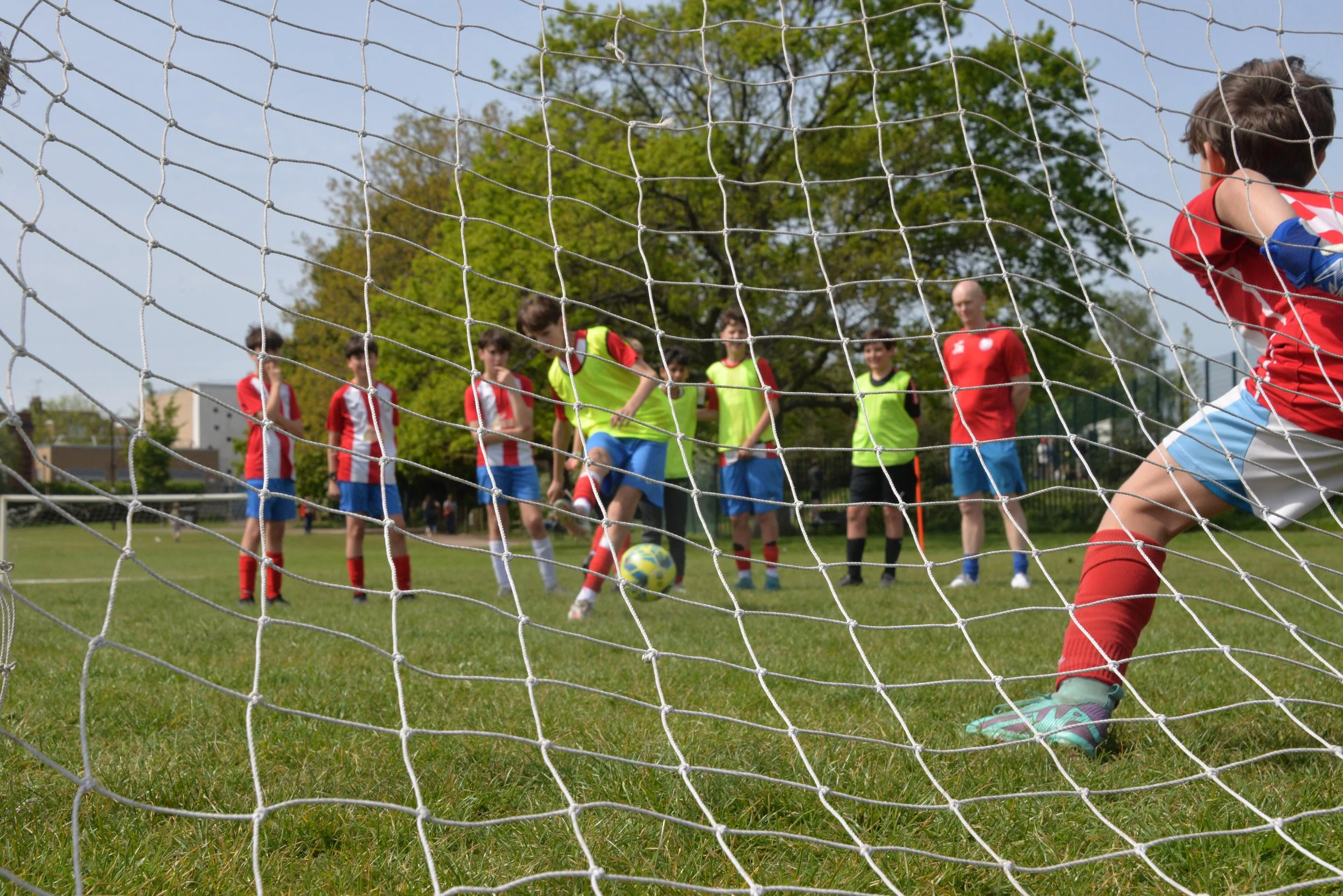Young soccer players practicing on a grassy field, seen from behind the goal net, with a focus on a child in red shorts and a red jersey taking a shot at the goal, while other players and a coach watch in the background.