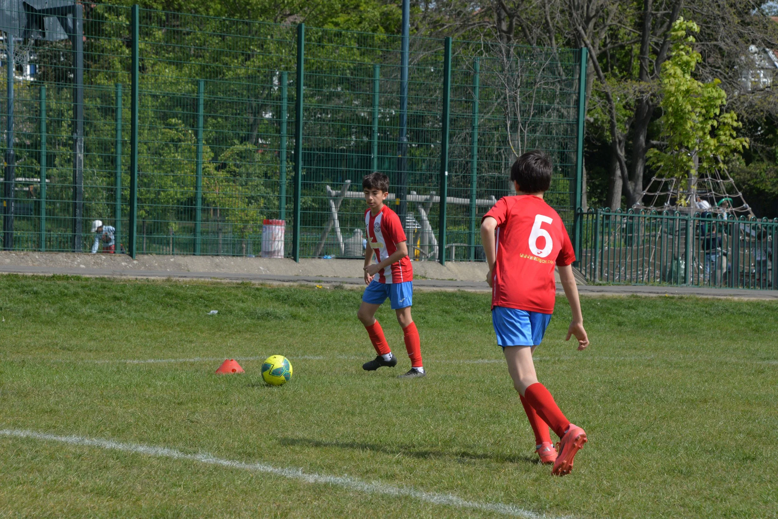 Two young boys playing soccer on a grassy field, one wearing a red and white striped jersey and the other in a solid red jersey with the number 6, with trees and a playground fence in the background.