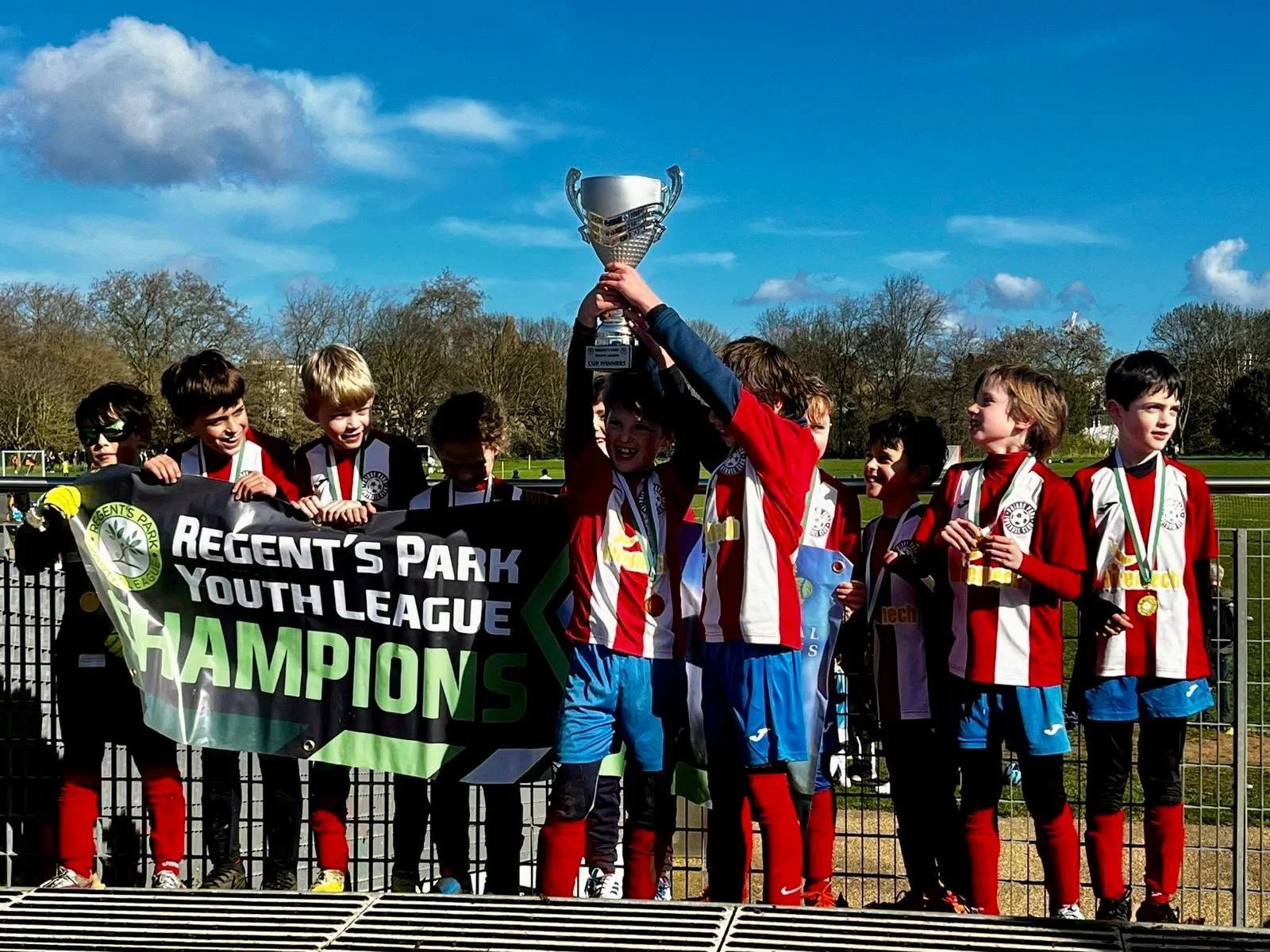 Young boys in red and white sports uniforms celebrating as they hold a trophy and a large banner that reads 'Regent's Park Youth League Champions' on a sunny day in a park.