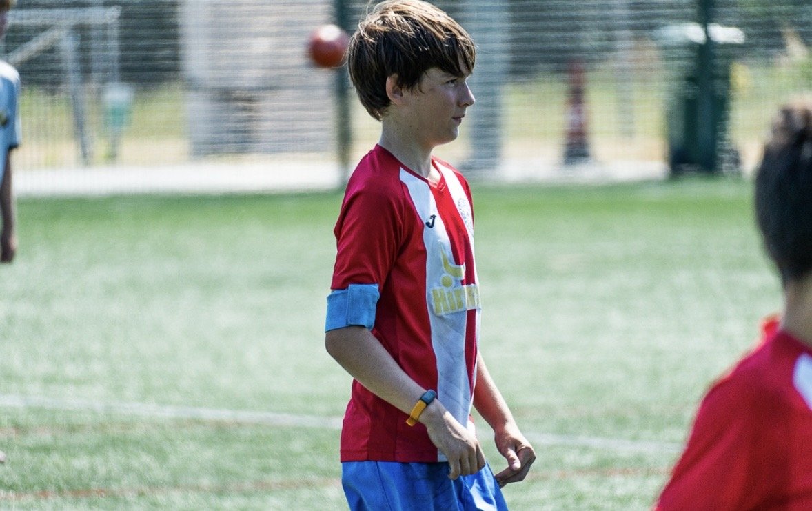 A young boy standing on a soccer field wearing a red and white jersey, blue shorts, and a yellow wristband, with a blurred background of a fence and other players.