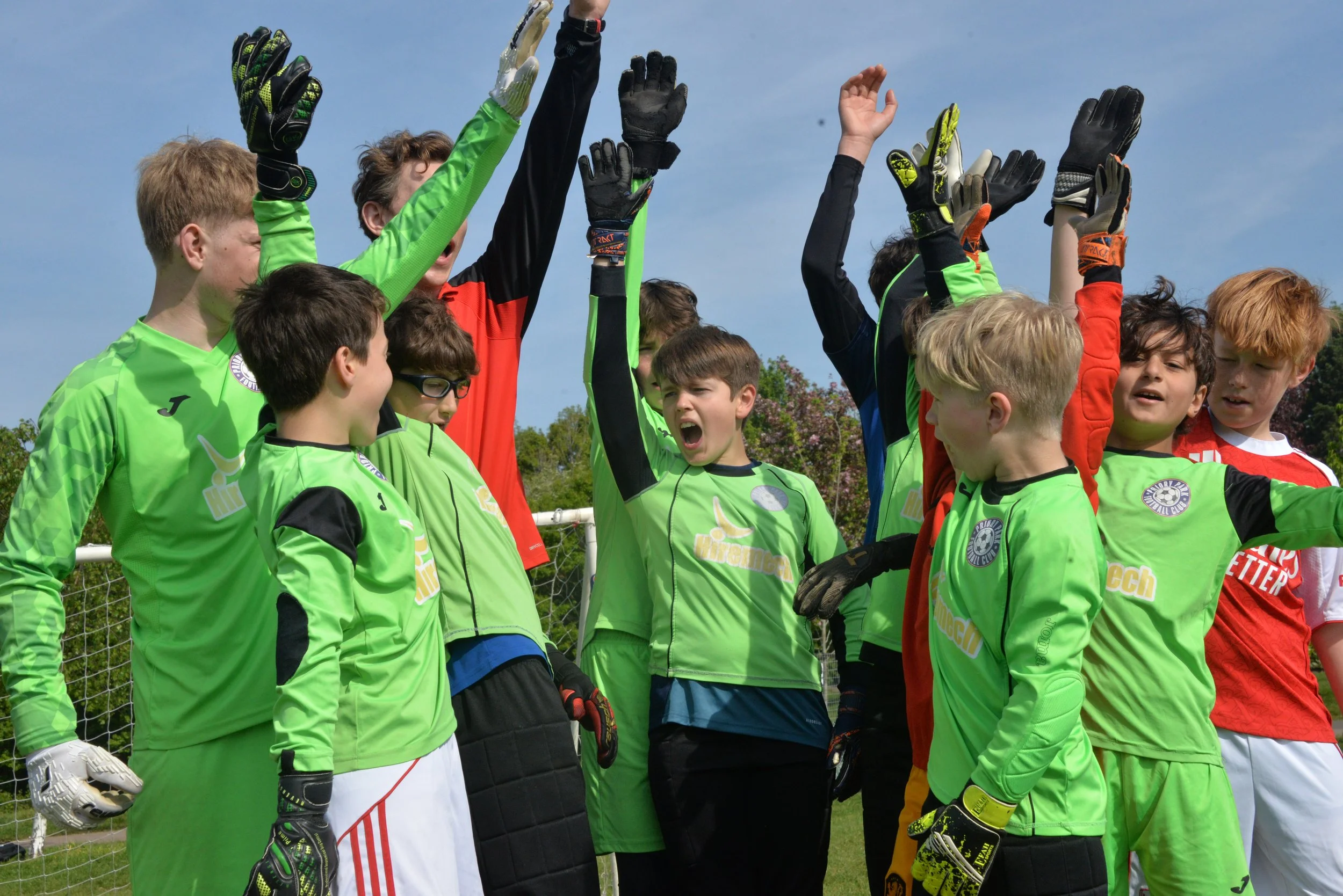 A group of young soccer players, including goalkeepers in green jerseys, raising their hands in a huddle on the field during daytime with a blue sky and trees in the background.