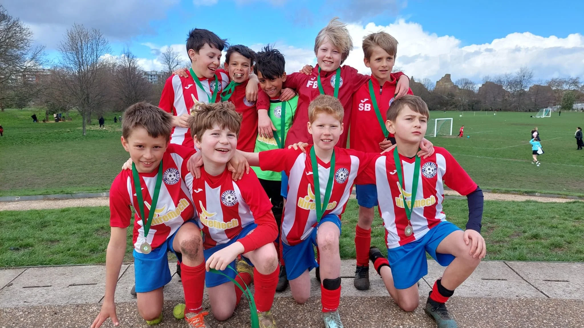 Group of young boys in soccer uniforms with medals, kneeling and standing on a grassy field after a soccer game, with other children playing in the background.