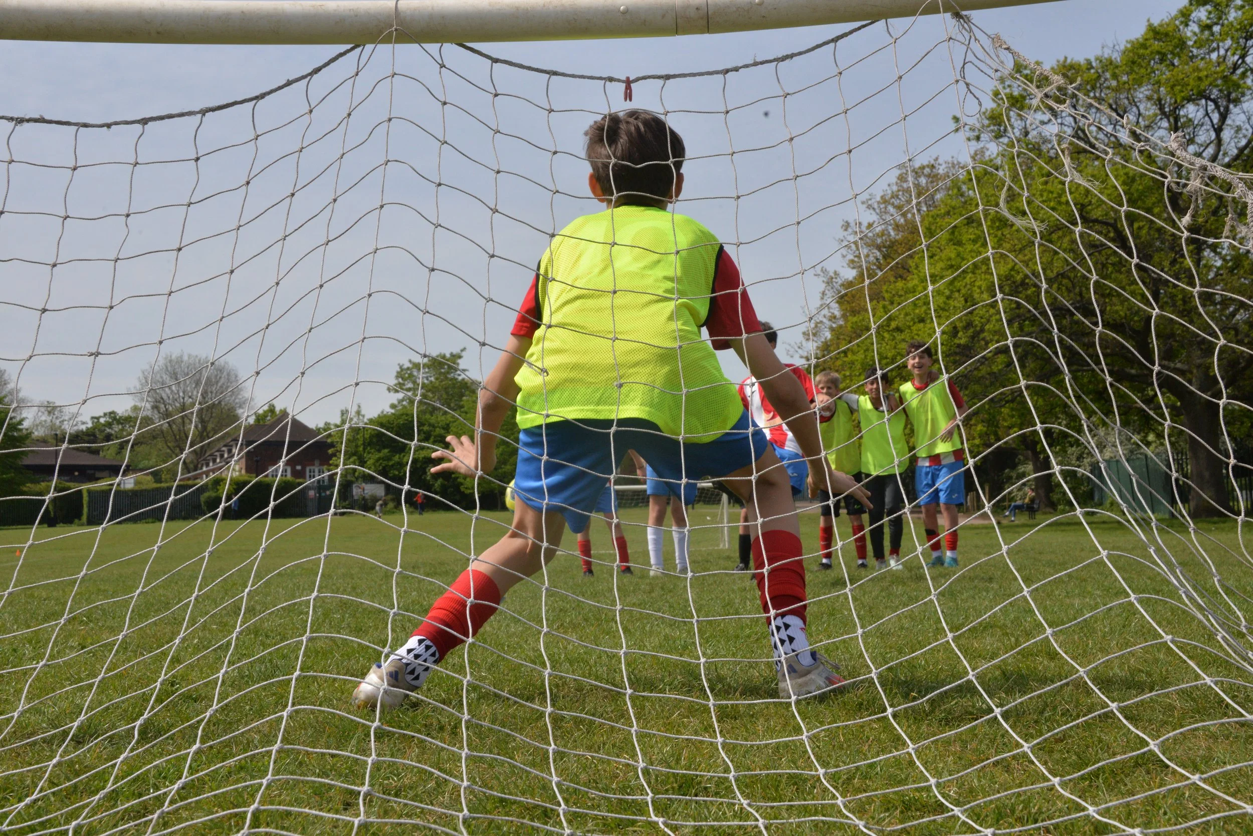 Children playing soccer on a field as seen through the goal net from behind the goalkeeper, with players in bright vests and trees in the background.