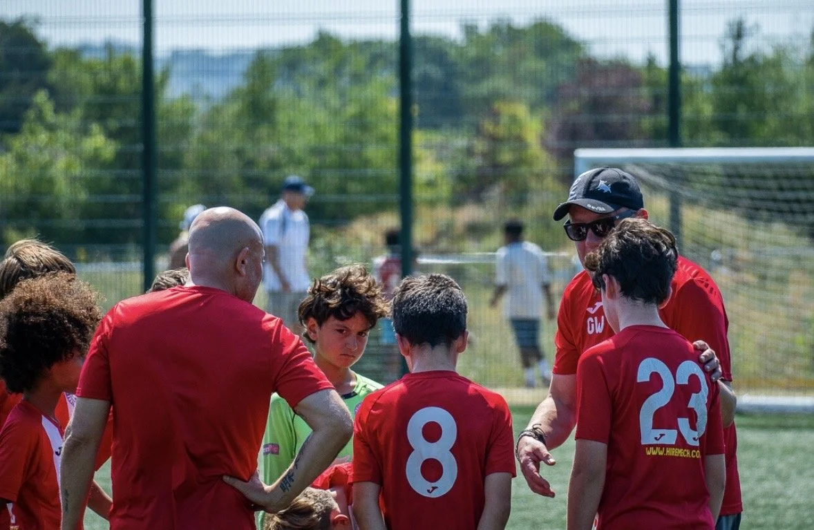 A youth soccer team gathered around their coach during a game on a sunny day, with a grassy field and goal in the background.