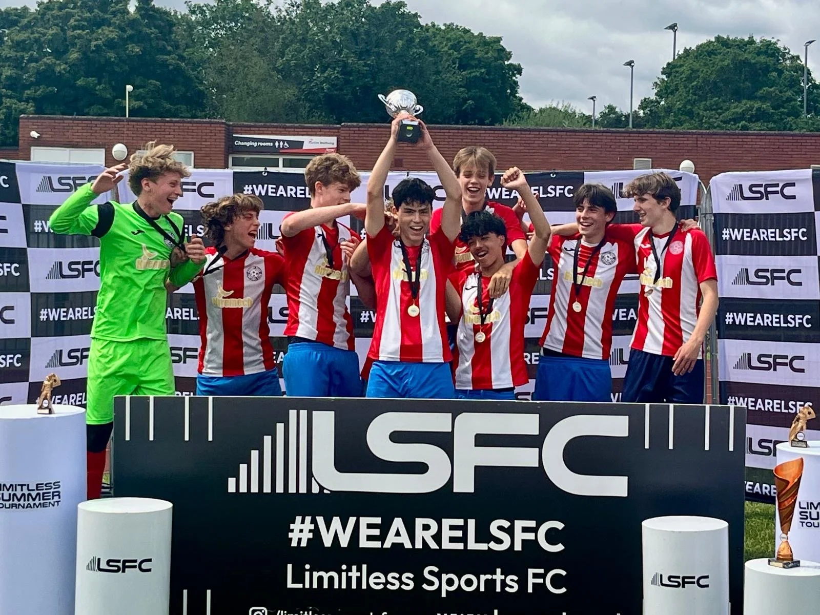Group of young soccer players celebrating their victory on a podium with medals and a trophy, wearing red and white striped jerseys and blue shorts, behind a black banner with the hashtag #WEARELSFC and the text 'Limitless Sports FC', outdoors with cloudy sky and trees in the background.