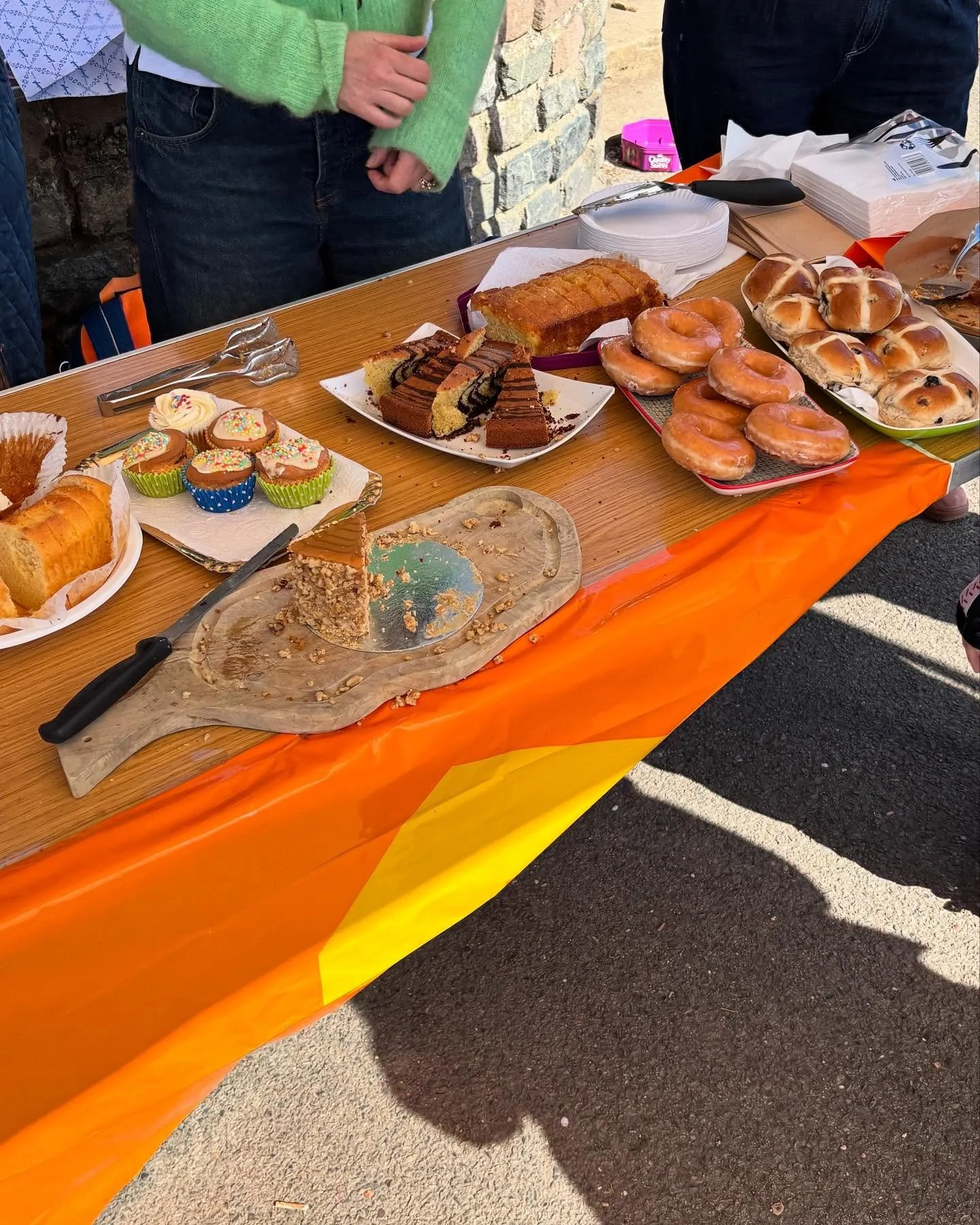 Table with assorted baked goods, including cupcakes with colorful sprinkles, slices of cake, a loaf cake, donuts, and bread rolls displayed at an outdoor event.