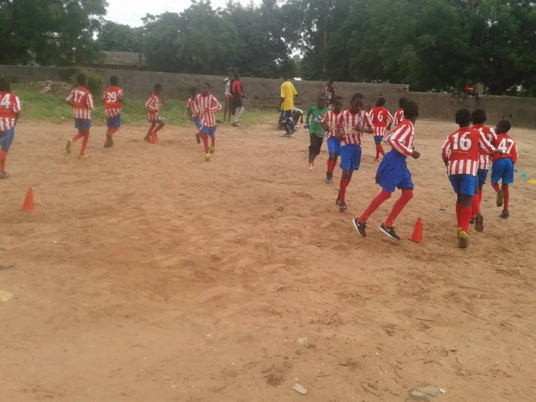 Group of young children in red and white striped soccer uniforms running on a dirt field with orange cones set up, likely during a training or practice session in an outdoor setting with trees in the background.