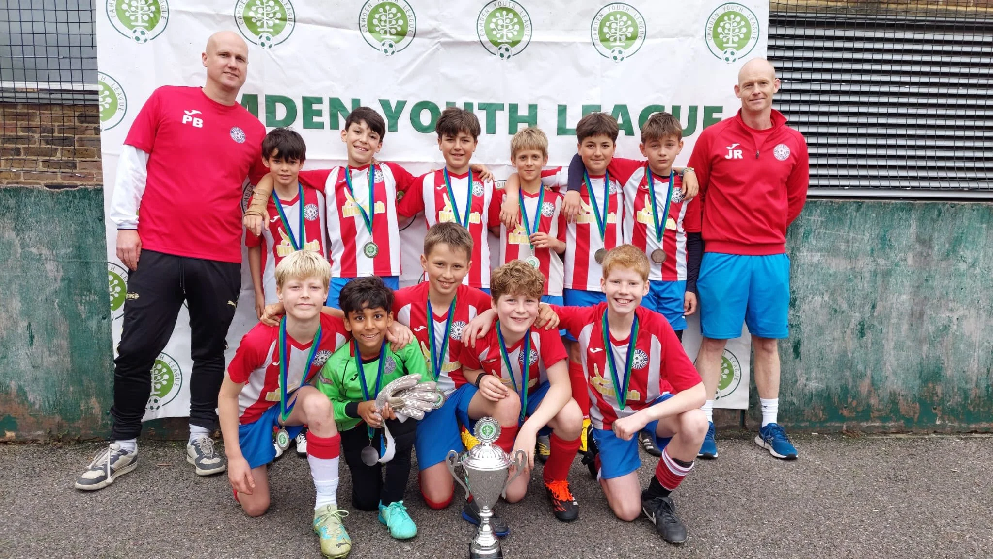 A youth soccer team of boys and their coaches posing for a photo after winning a tournament, wearing red and white striped jerseys, medals around their necks, with a trophy placed on the ground in front of them, in front of a banner that reads 'UNDER