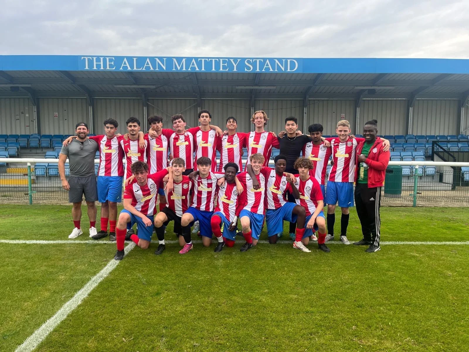 Group of young soccer players and coaches posing for a team photo on a soccer field, with a stand labeled 'The Alan Matty Stand' in the background.