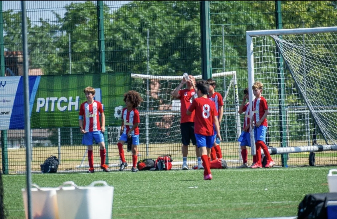 Young soccer players in red and white jerseys on the sideline of a soccer field, with some drinking water and waiting to play.
