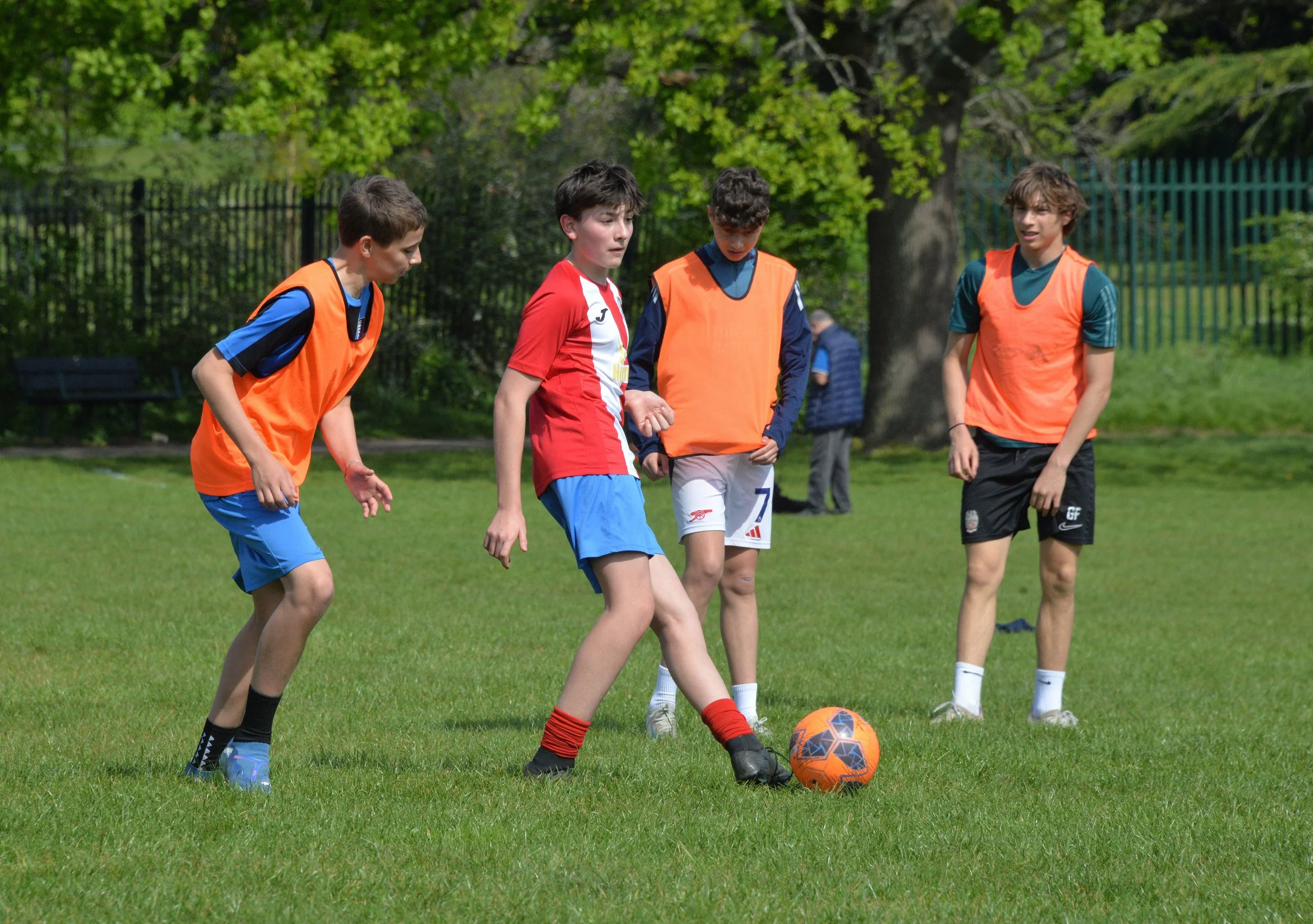 Four boys are playing soccer on a grassy field. Two of them are wearing orange pinnies, one is in a red and white jersey, and one in a teal shirt. They are focused on an orange and black soccer ball, with trees and a fence in the background.