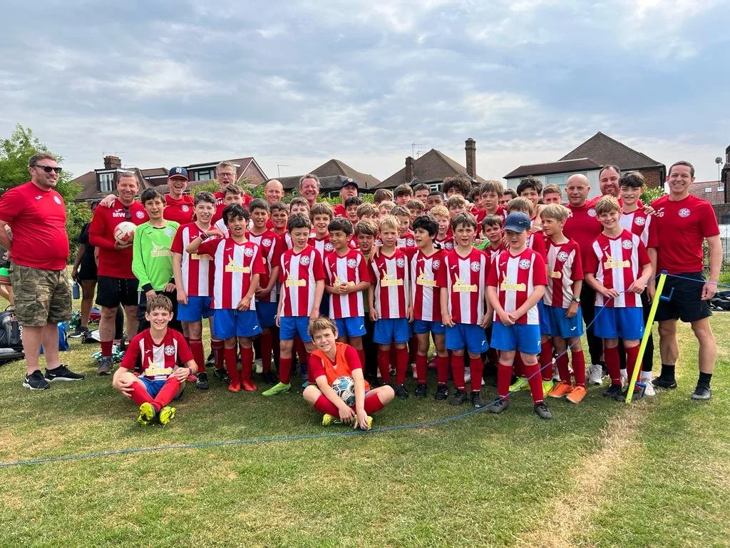 A group of young boys in red and white soccer uniforms with blue shorts, along with coaches and staff in red shirts, gathered on a grassy field for a team photo. There are houses and a cloudy sky in the background.