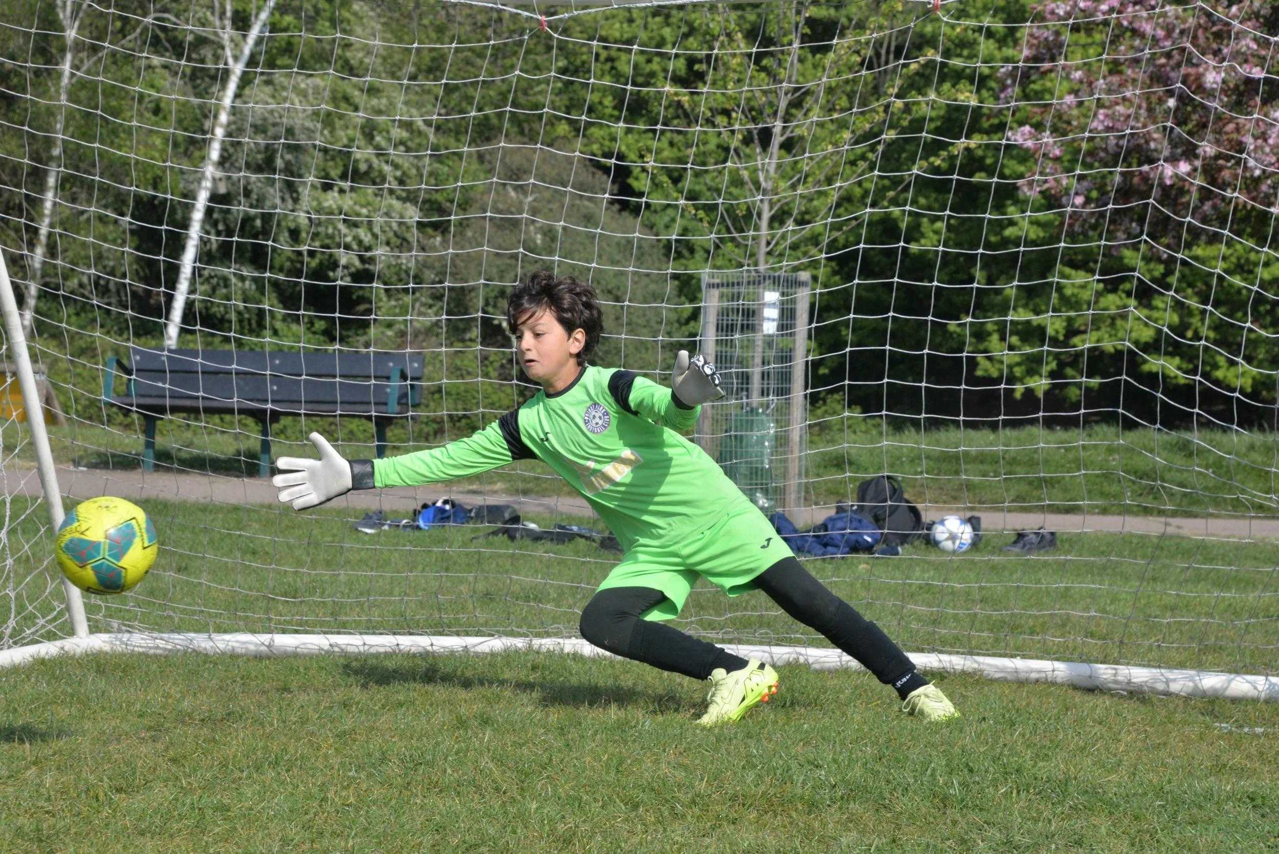 A young male soccer goalkeeper in a bright green uniform and gloves making a save during a match on a field with trees in the background.