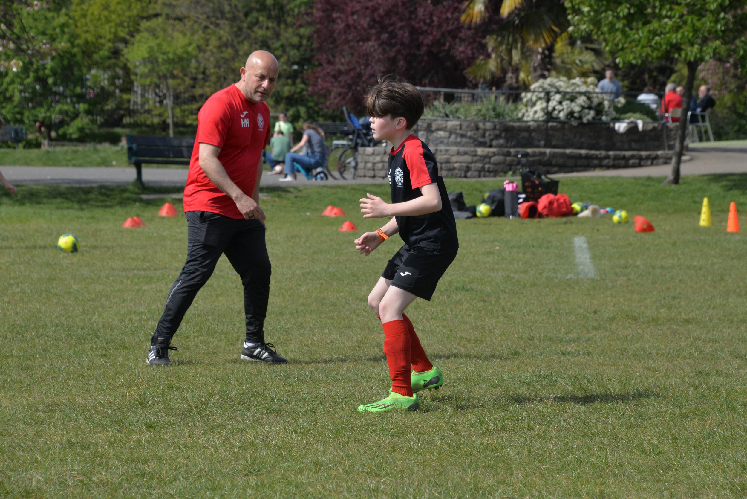 A man and a boy practicing soccer on a grassy field surrounded by trees, with orange cones and tennis balls scattered on the ground.