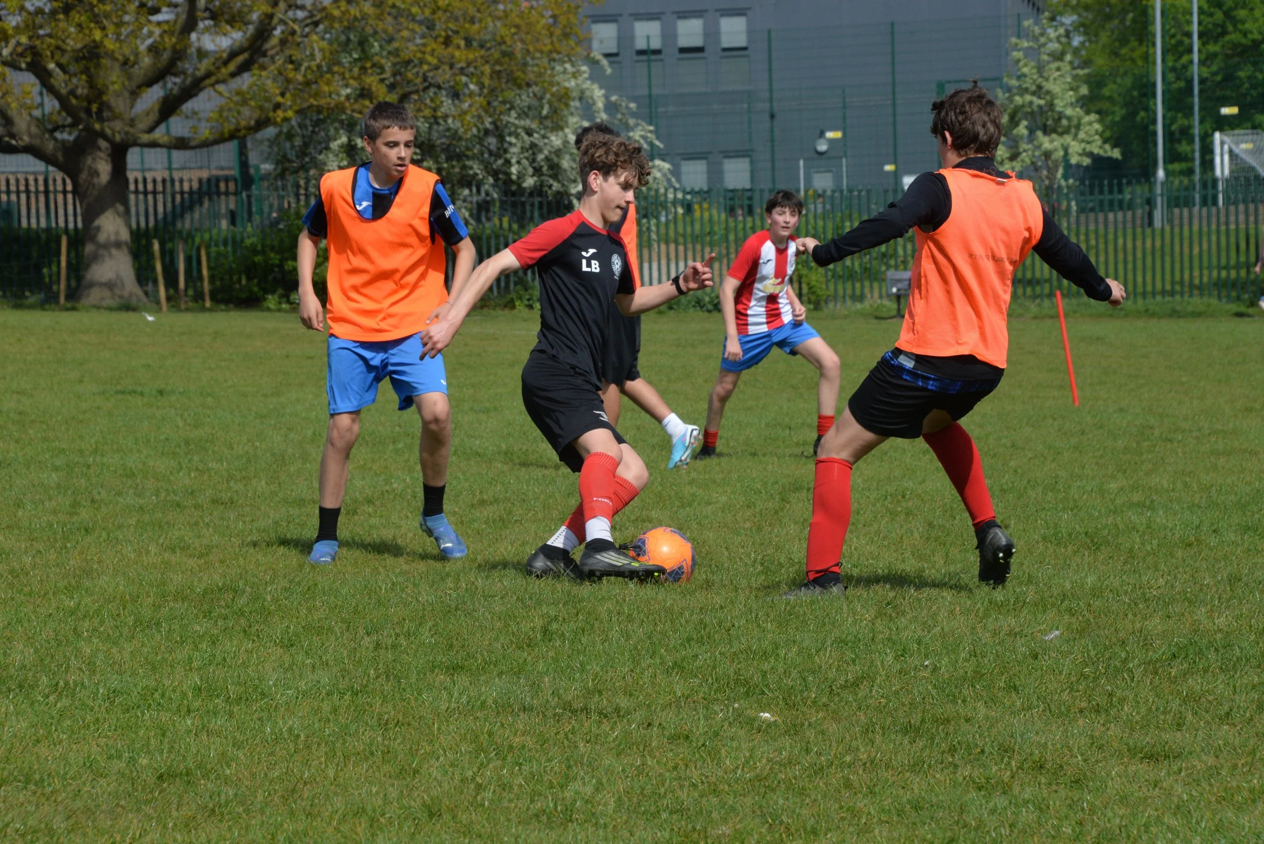 Four boys playing soccer on a grassy field, with two in orange vests, one in black, and one in a red and white striped jersey, surrounded by trees and a fence.