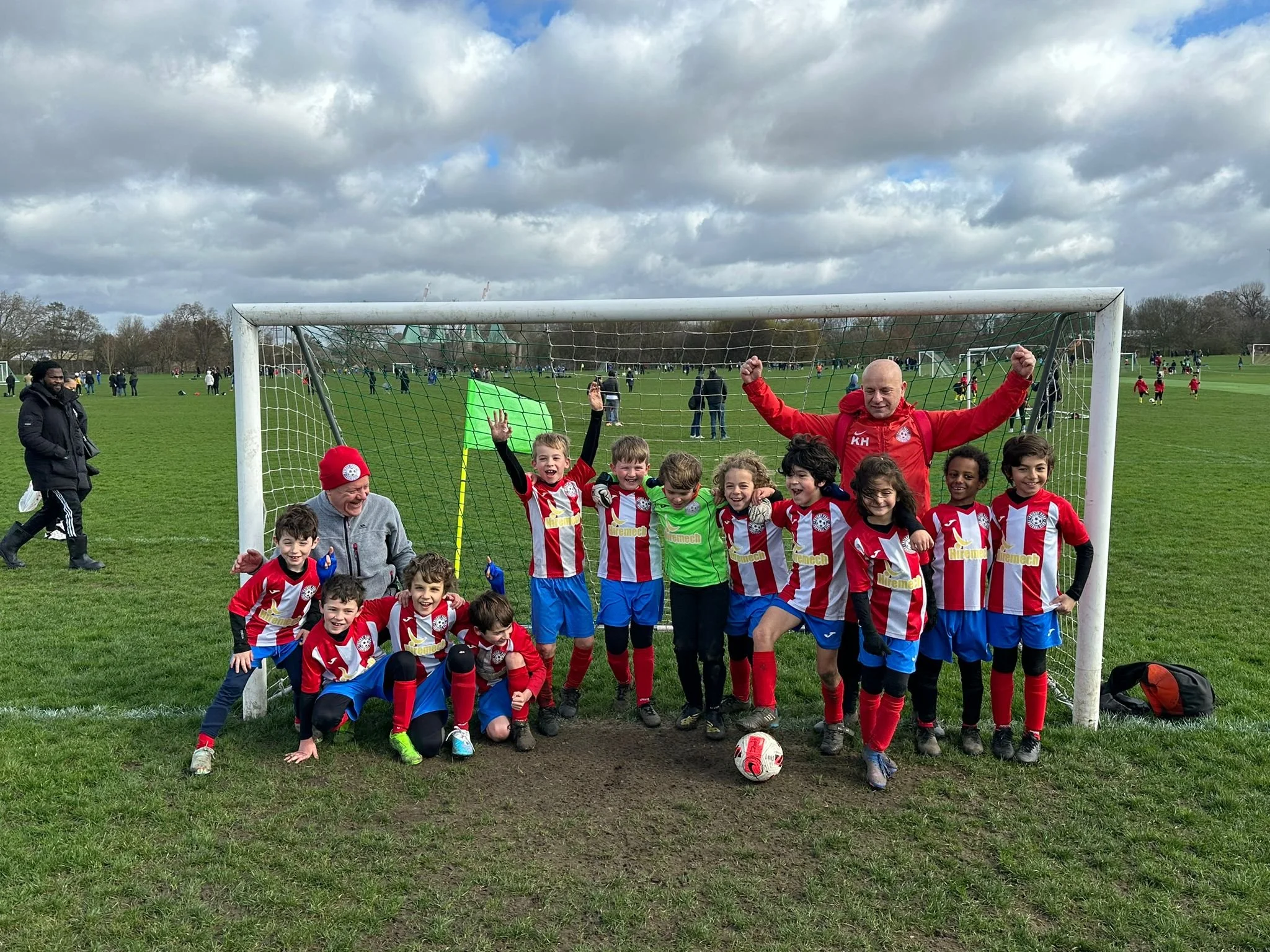 A children's soccer team with their coach posing in front of a goalpost on a grassy field. The children are wearing red and white striped uniforms, and the coach is wearing a red jacket. Some players are kneeling, and others are standing, smiling wit