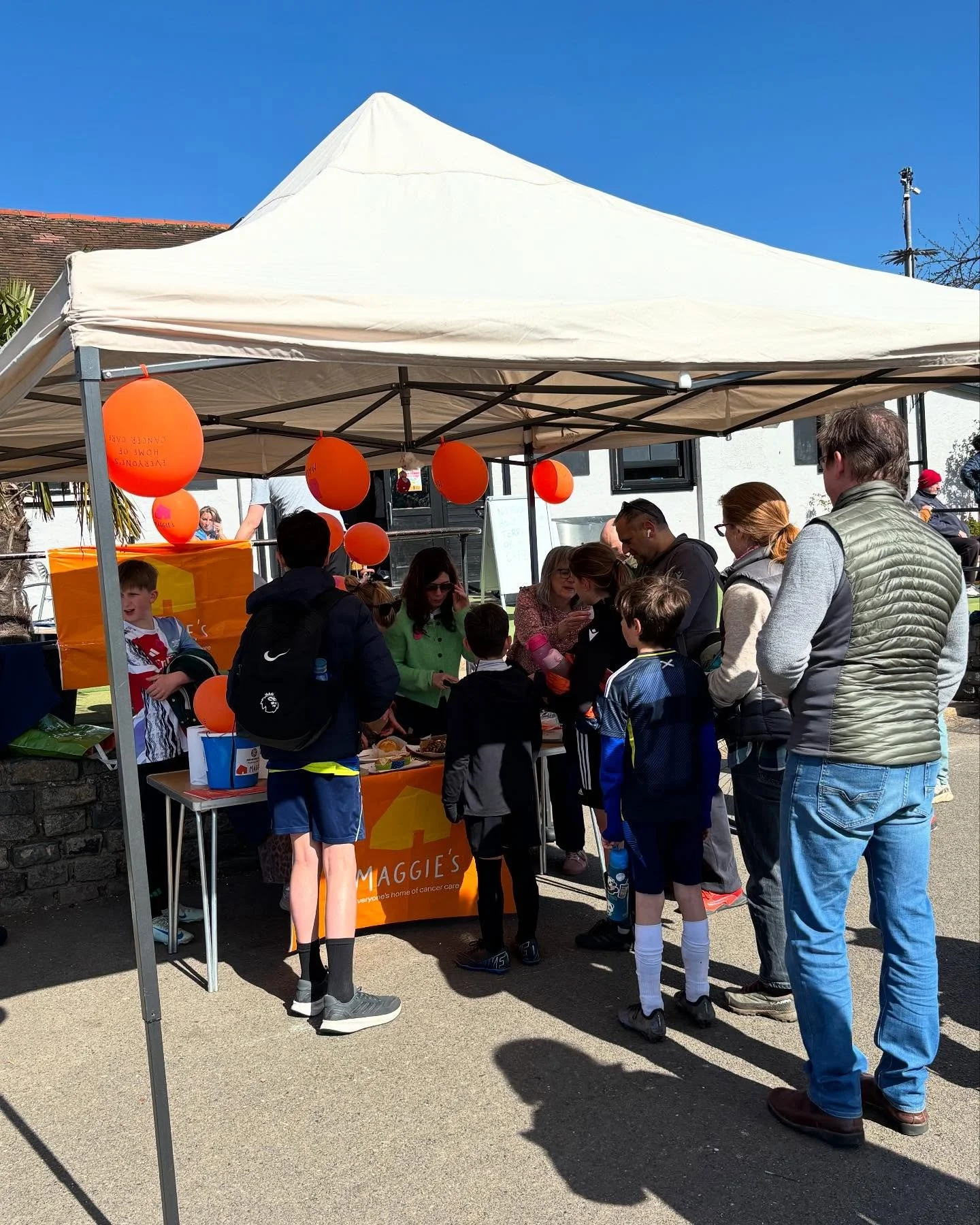 People gathered under a white canopy tent at an outdoor event, with orange balloons and a table displaying information or items, as part of a community or charity event.