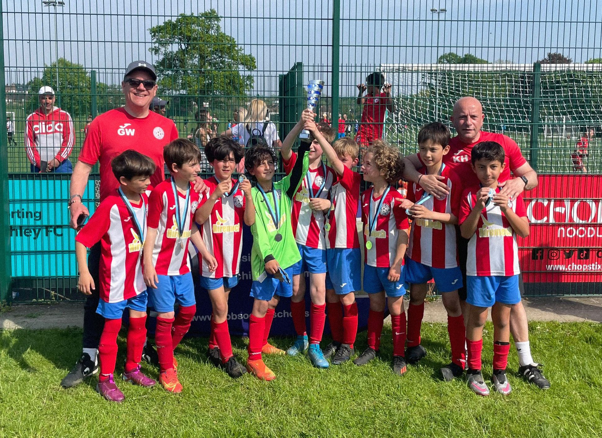 A group of young soccer players in red and blue uniforms celebrating after a match, with a coach and a trophy.